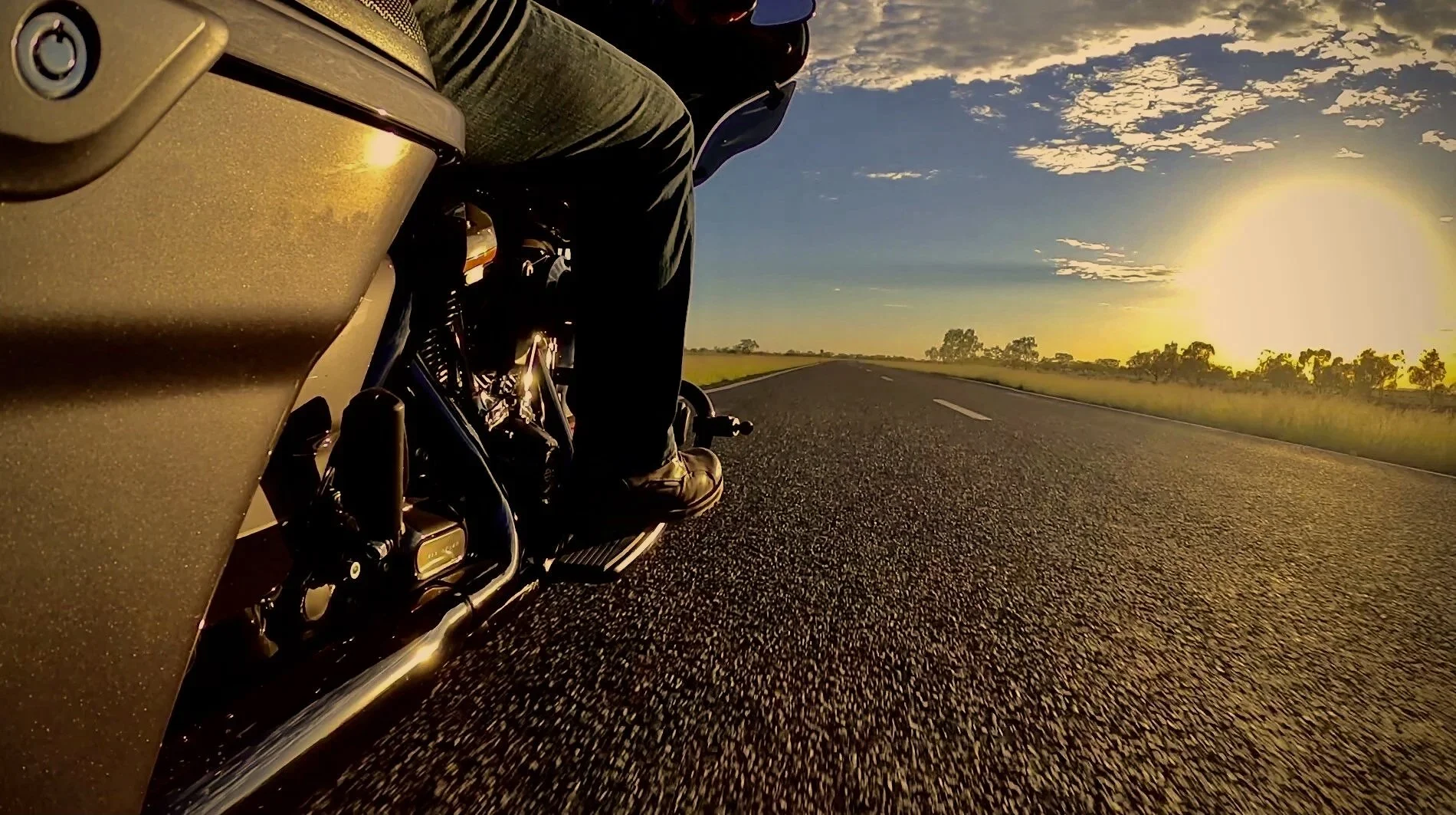 View of a motorcycle riding on an open road during sunset, capturing the side and engine details with a scenic landscape of trees and clouds in the background.