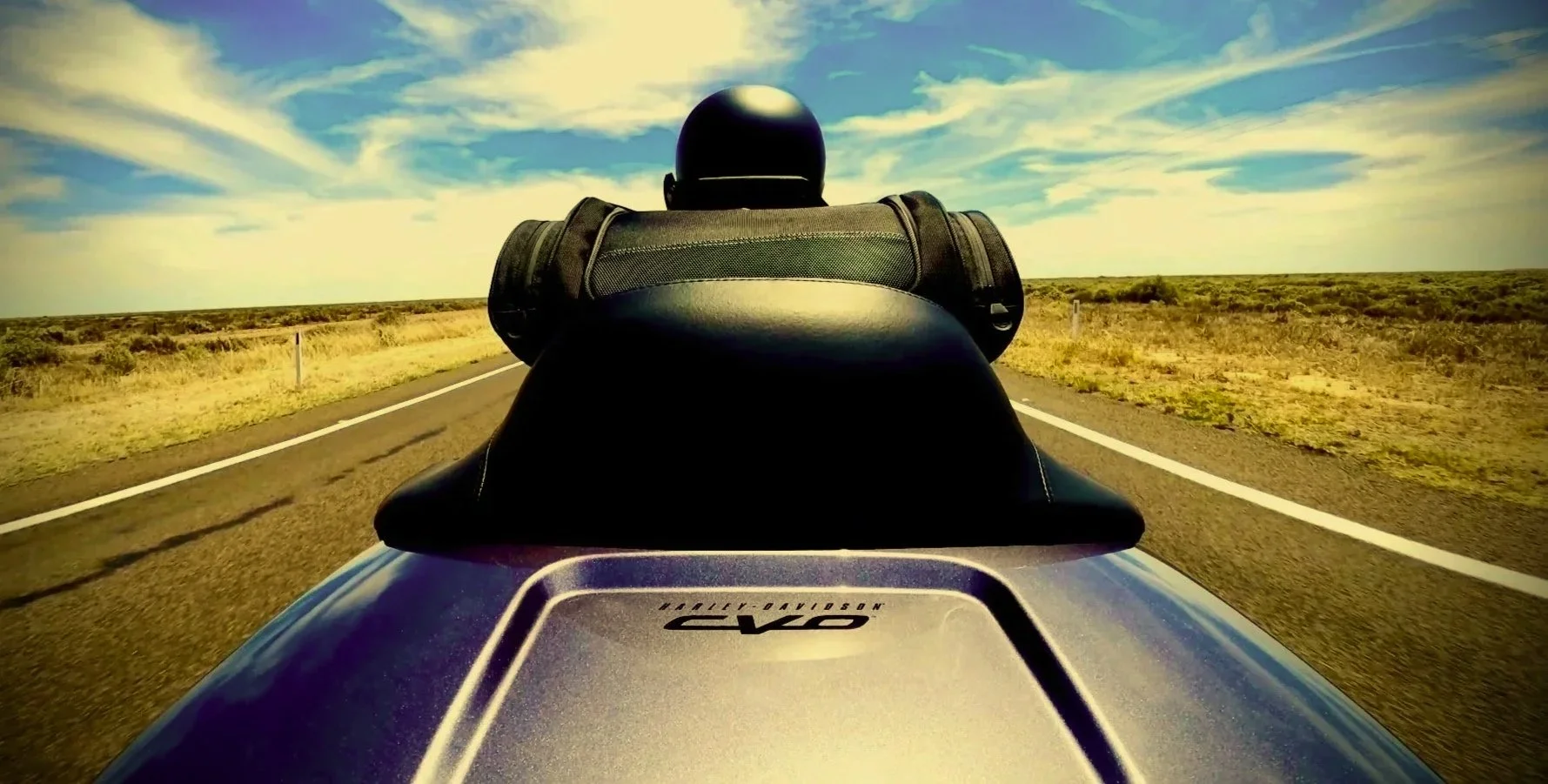 Person riding a motorcycle on an open desert road with a clear blue sky and wispy clouds.