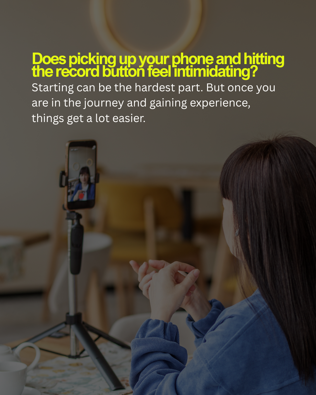 A woman with long dark hair in a blue top sitting at a table, recording herself with a smartphone on a tripod for a video or TikTok, with a message about overcoming intimidation when recording.