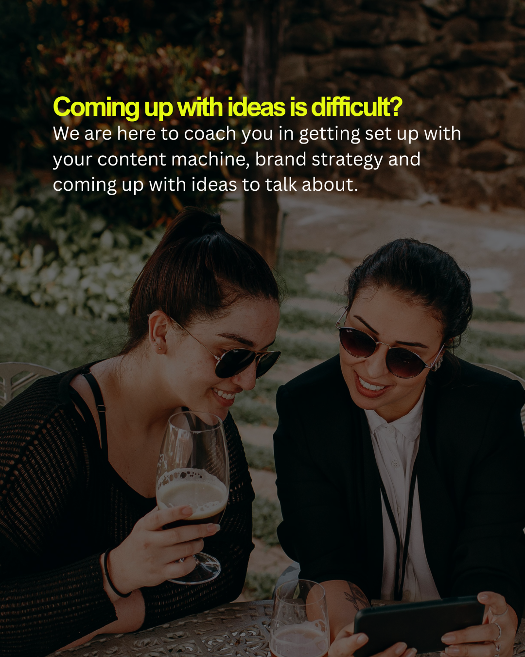 Two women with dark hair and sunglasses sitting at an outdoor table, smiling and looking at a smartphone while holding glasses of beer.