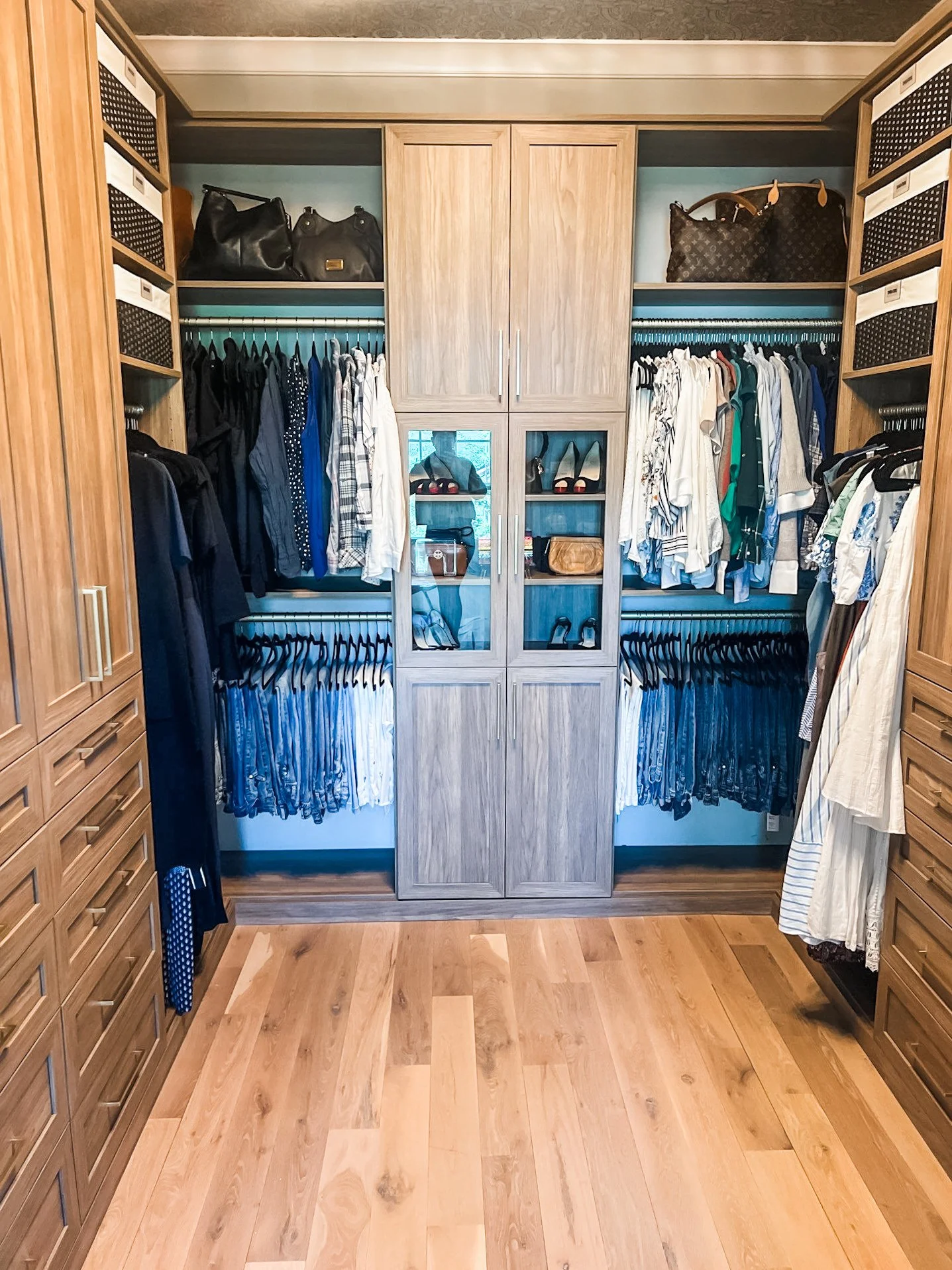 A walk-in closet with wooden shelves and drawers. Clothing hanging on rods, bags on upper shelves, and shoes on lower shelves behind glass doors.