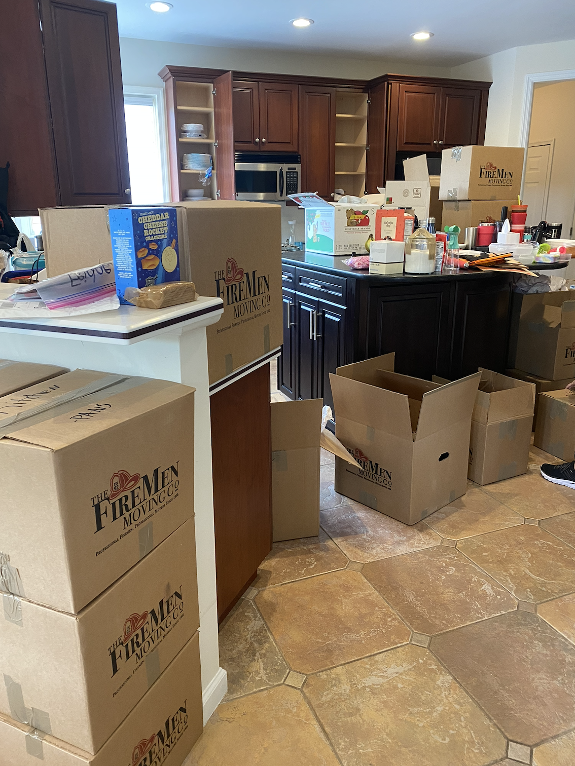 Kitchen filled with packed moving boxes labeled 'The Firemen Moving Co.' and various household items, with dark wood cabinets and a tile floor.