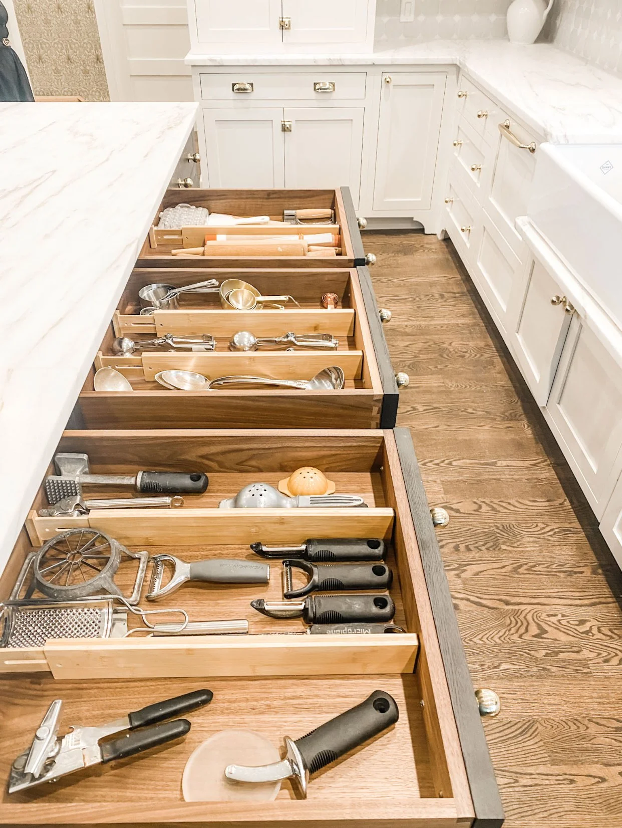 Kitchen with open wooden drawer containing various kitchen tools and utensils, white cabinets, and a marble countertop.