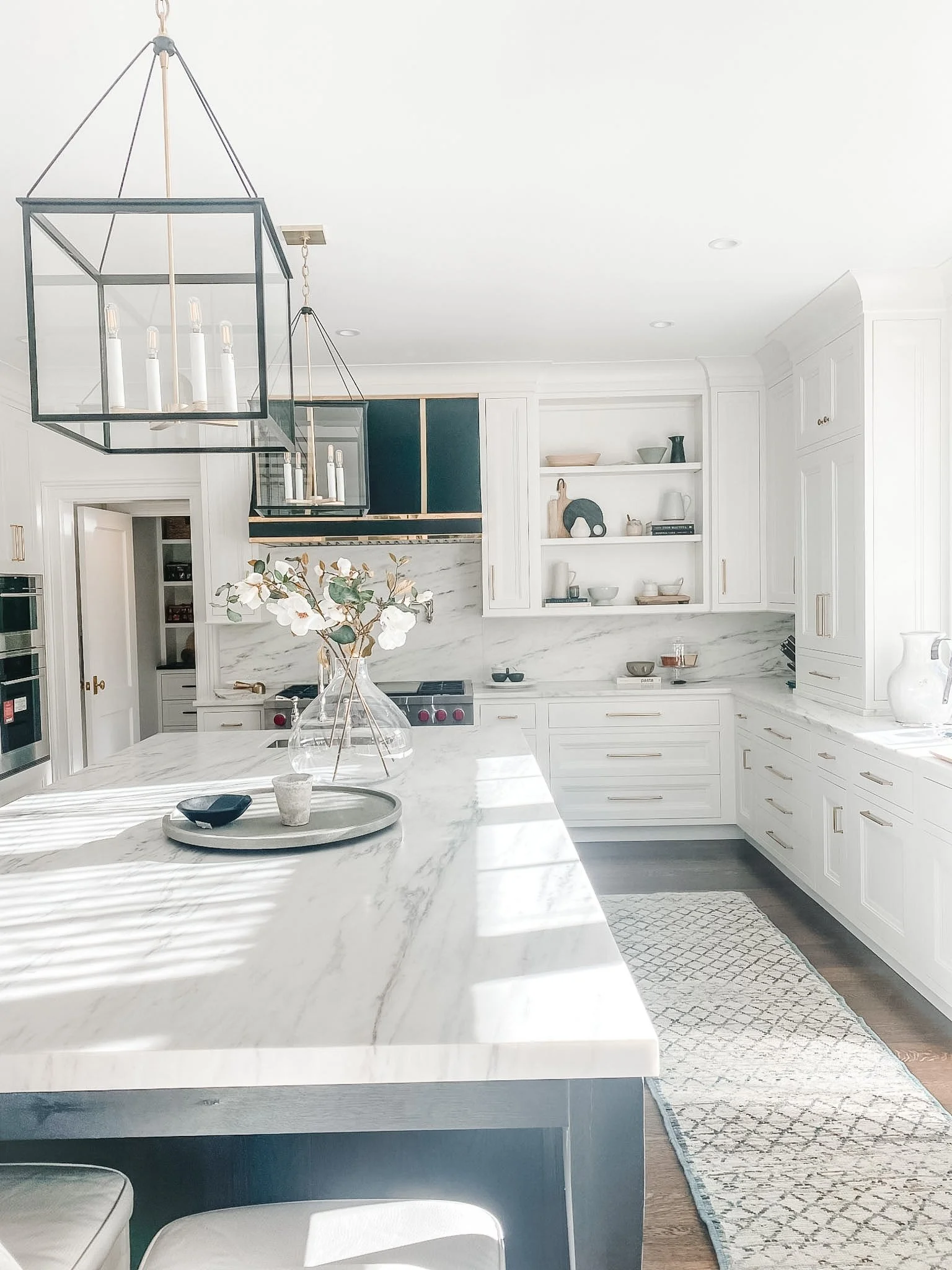 A modern kitchen with white cabinets, marble countertops, open shelves with dishes, a flower vase on the island, and a chandelier hanging from the ceiling.