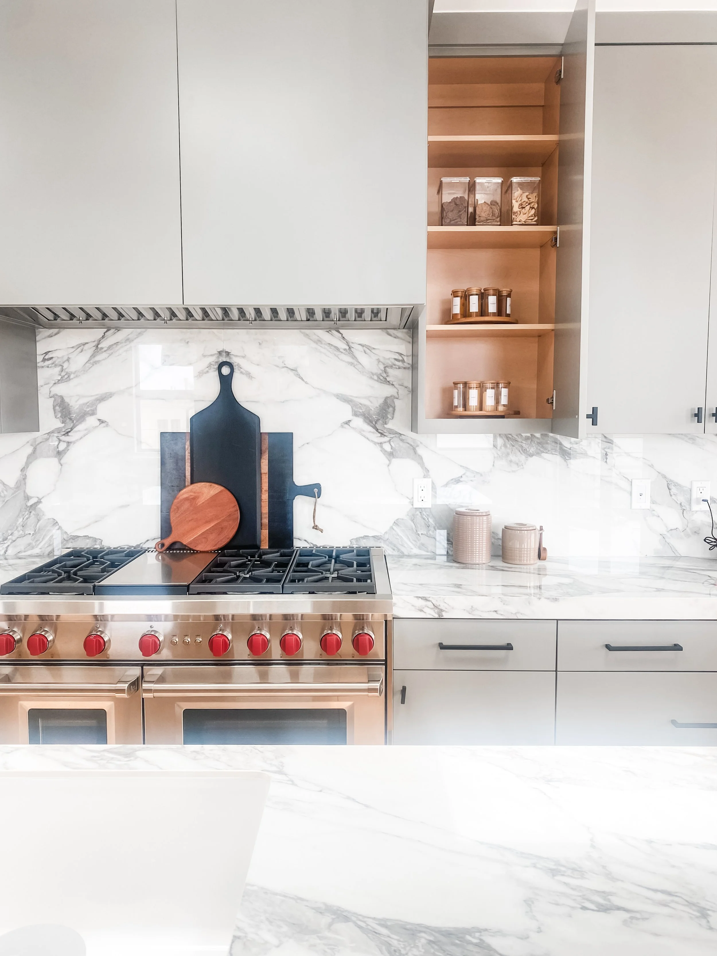 Modern kitchen with white cabinetry and marble countertops, stainless steel gas stove with red controls, open wooden cabinets with jars on shelves, and a marble backsplash.