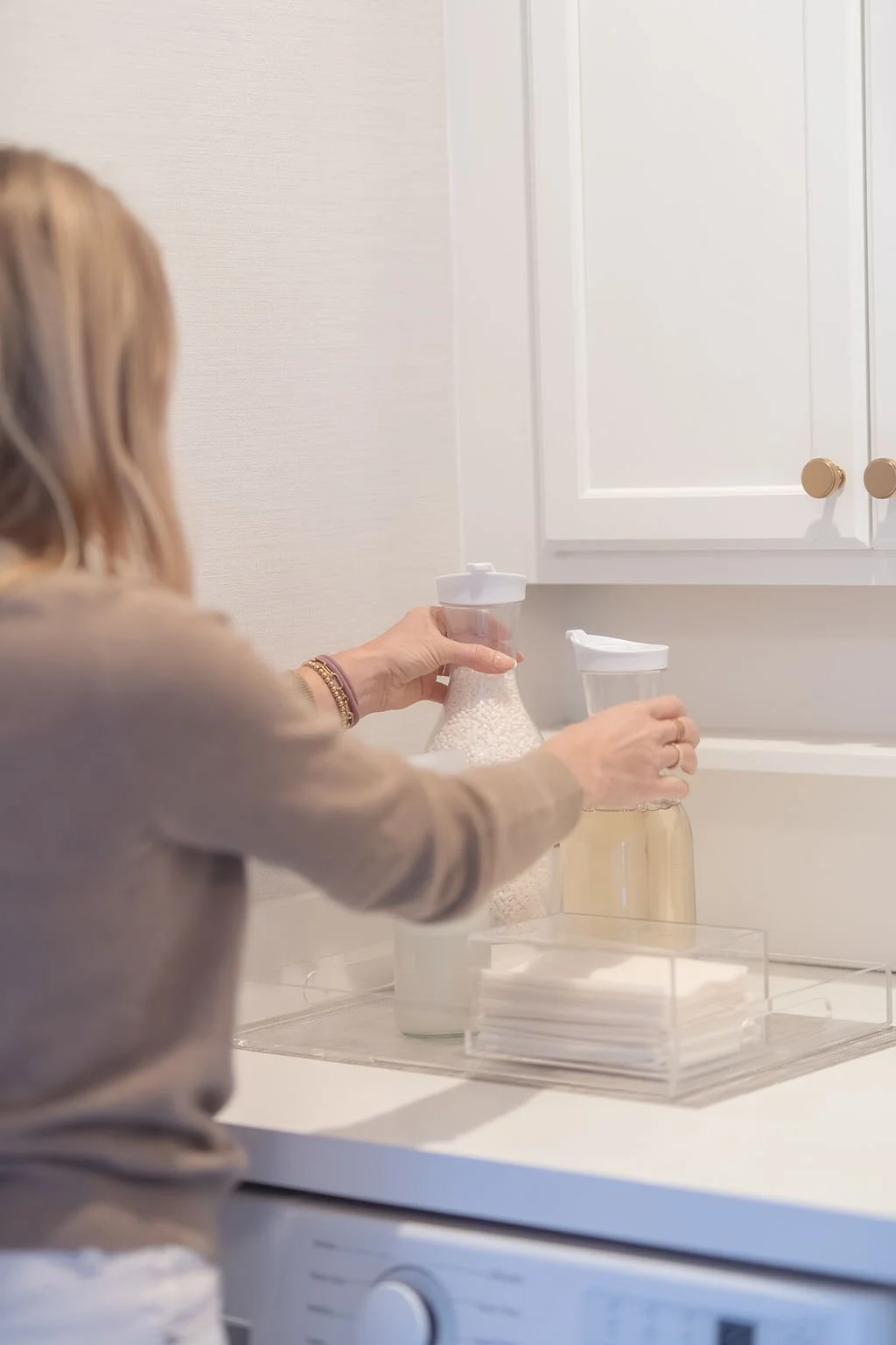 A woman in a beige sweater preparing bottles for a baby in a white kitchen with white cabinets and a neutral wall.