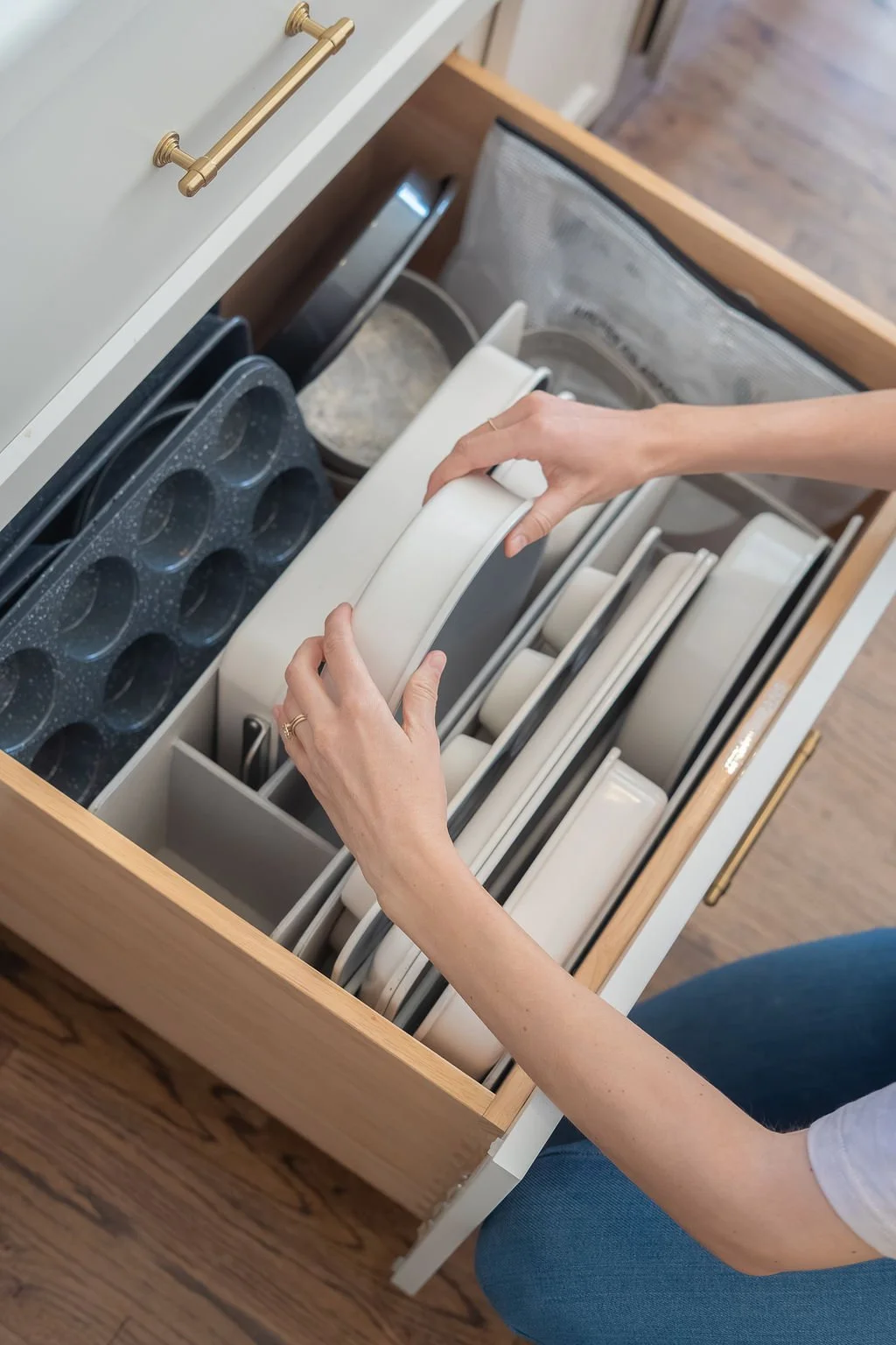 Person organizing white plates in a kitchen drawer filled with dishware, including baking pans and bowls.