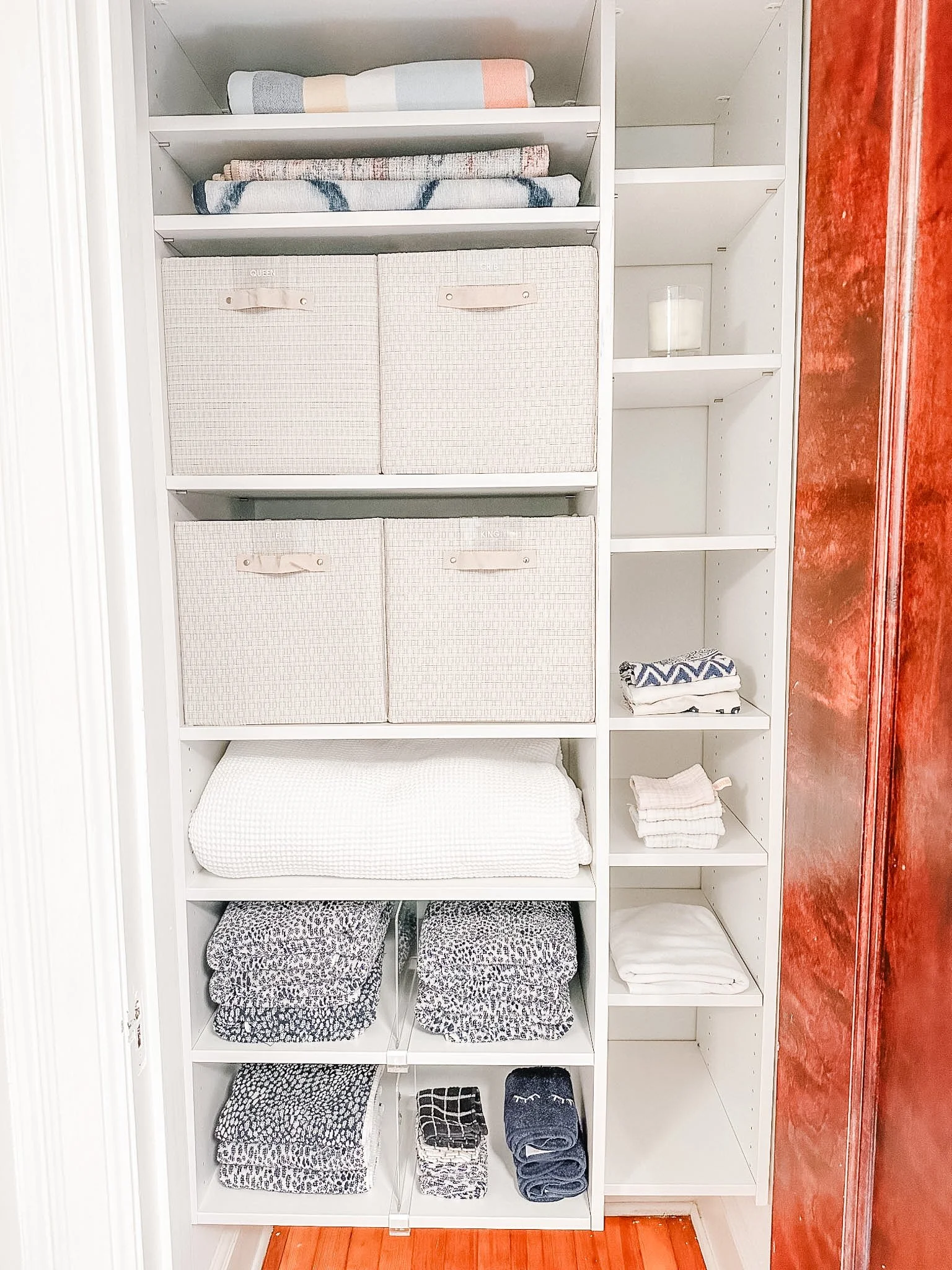 A white bookshelf with foldable fabric storage bins, folded towels, and various linens on the shelves, with a red spalsh wall on the side.