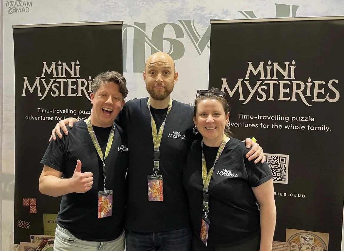 Three smiling people at a Mini Mysteries event, wearing black shirts with the logo. Two are giving a thumbs up, and they are standing in front of black banners with the Mini Mysteries logo and a QR code.