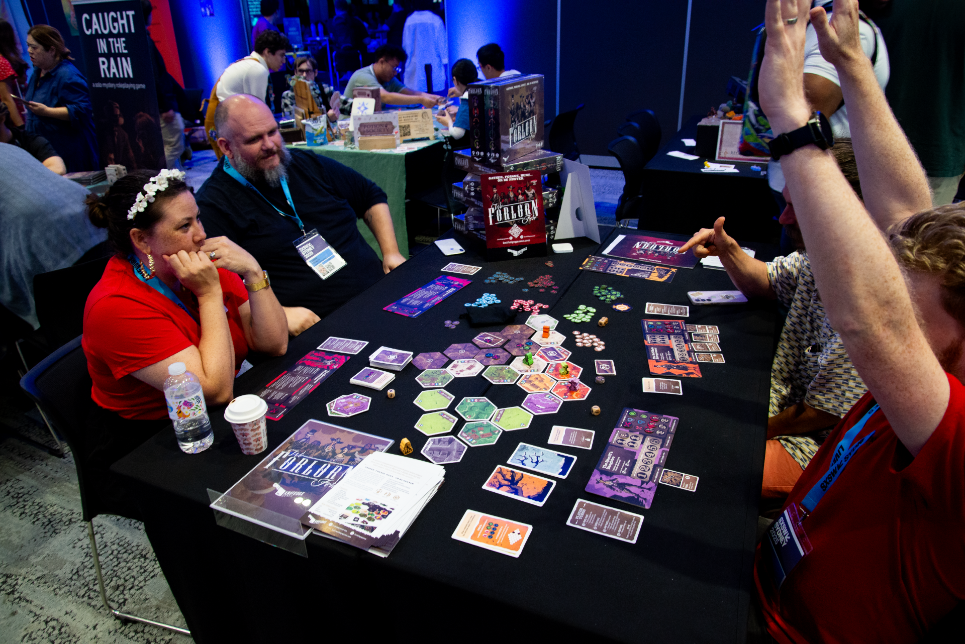 People playing a strategy board game called Forlorn at a convention. The game has hexagonal tiles, game cards, and tokens on the table. Several players and onlookers are visible, with other booths and attendees in the background.
