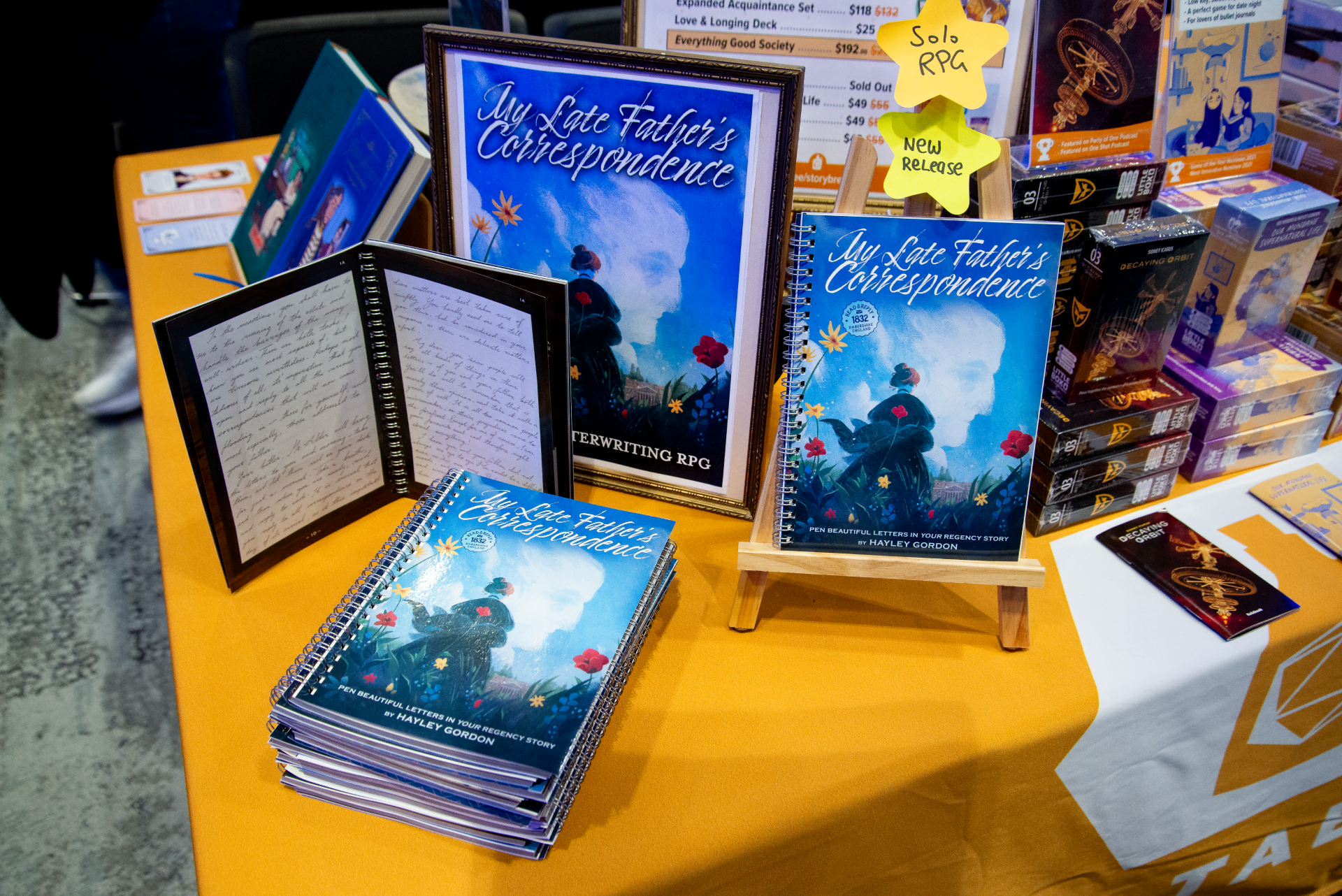 Display of a book titled "My Late Father's Correspondence" with a blue cover featuring an illustration of a woman in a field. Several copies of the book are stacked on a yellow table, with additional copies on a small wooden stand. Bright yellow sticky notes with handwritten labels "Solo RPA" and "New Release" are attached. In the background, there are other books and display materials on the table.