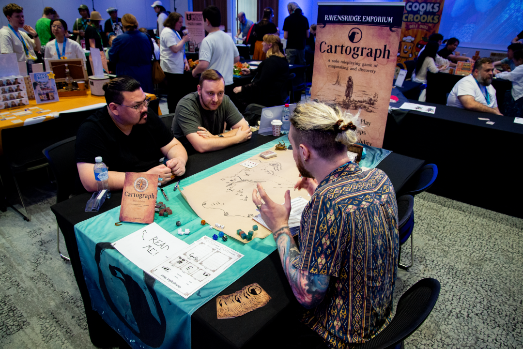 People playing the tabletop game 'Cartograph' at a convention or gaming event, with promotional banners and a variety of games on display in the background.