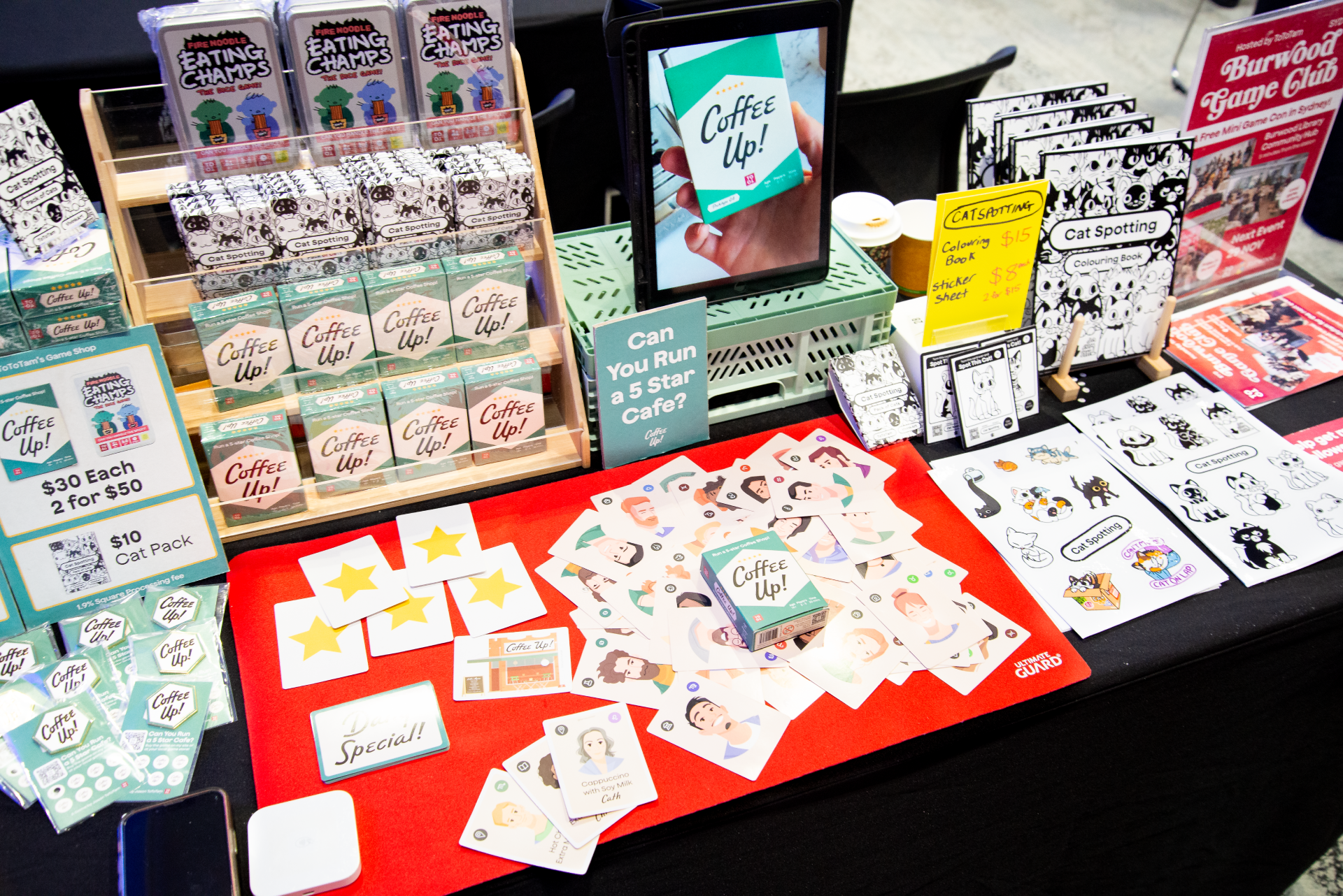 A tabletop display promoting various products related to cats and coffee. The display includes small toys, books, coloring books, stickers, cards, and a sign asking if you can run a five-star cafe. There are also snacks and a tablet showing a promotional message for coffee.