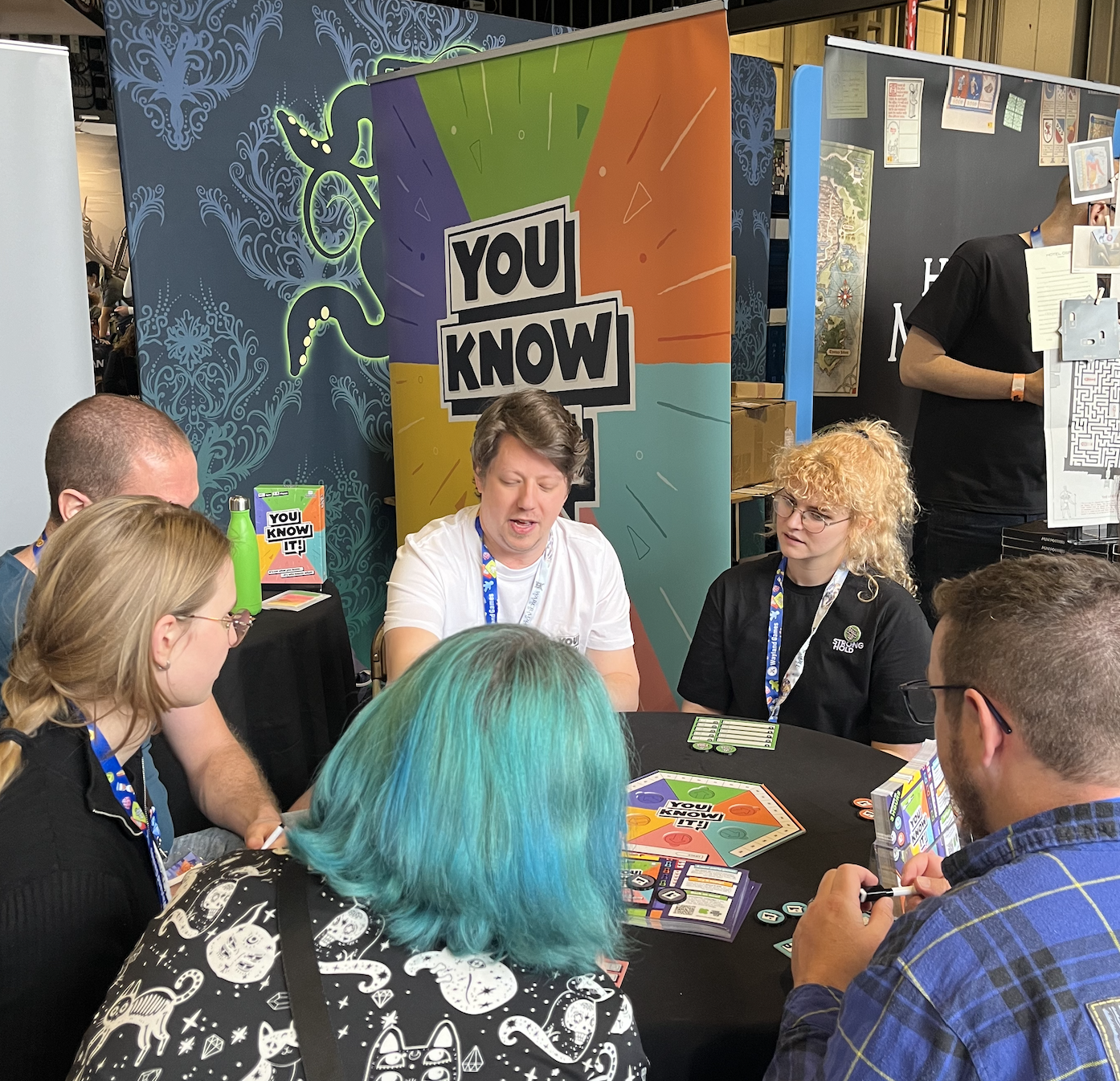 Group of people playing a board game called 'You Know It!' at a booth with a colorful banner.
