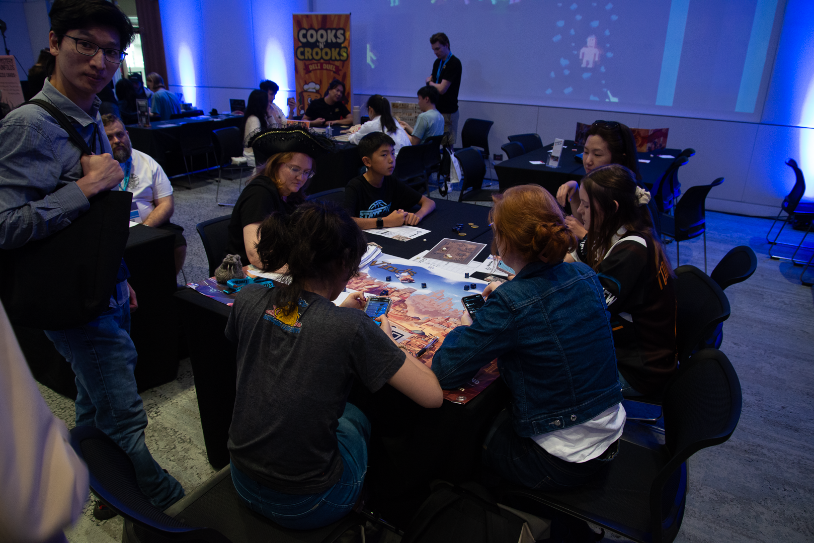 People playing a board game at a table during a gaming event or convention, with some participants using smartphones and a person dressed as a pirate, in a room with blue lighting and promotional banners.