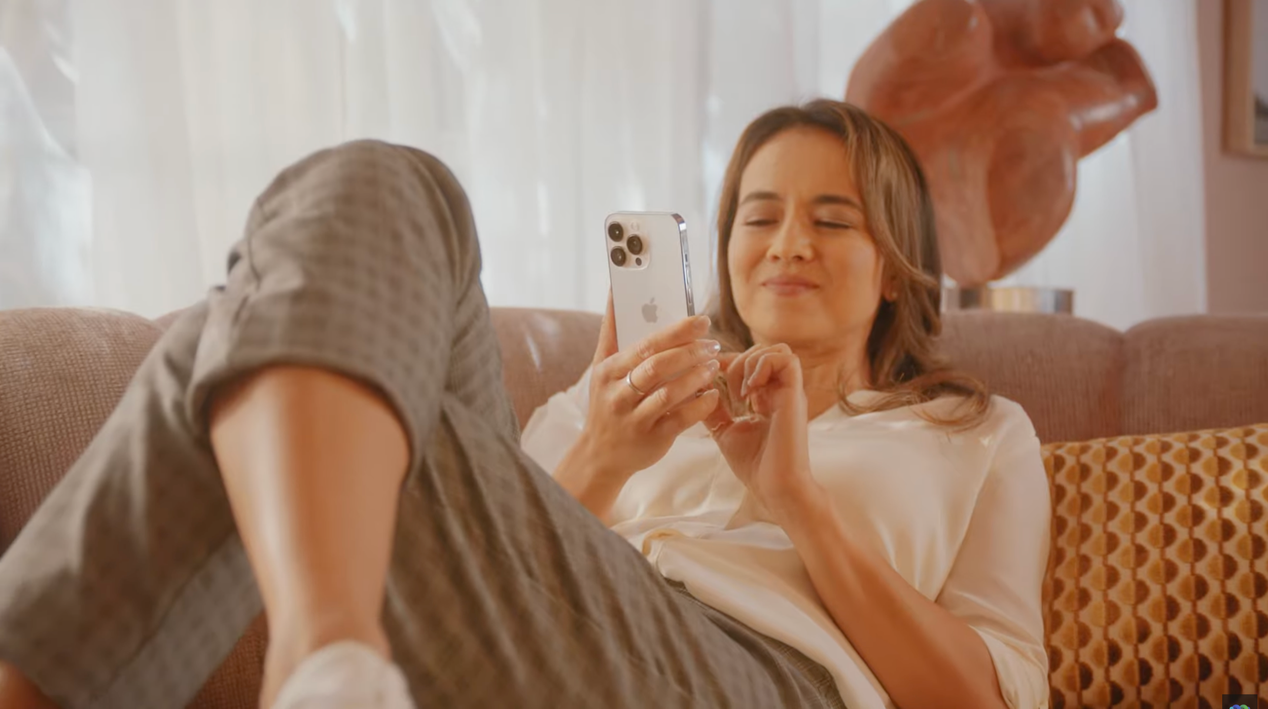 Woman in beige pants and cream blouse lounging on a sofa, holding an iPhone, with a large abstract sculpture in the background.