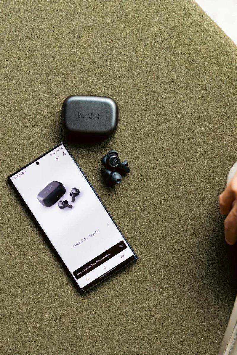 Product photography by ST Davis for Bang & Olufsen x Cisco — wireless earbuds with charging case and smartphone app on a textured surface