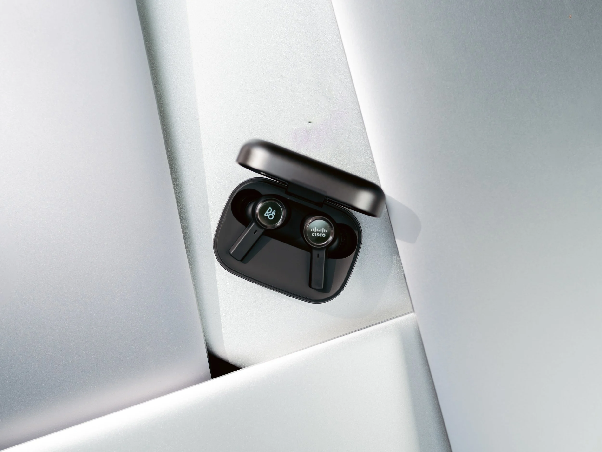 Product photography by ST Davis for Bang & Olufsen x Cisco — black wireless earbuds in open charging case on a geometric silver surface