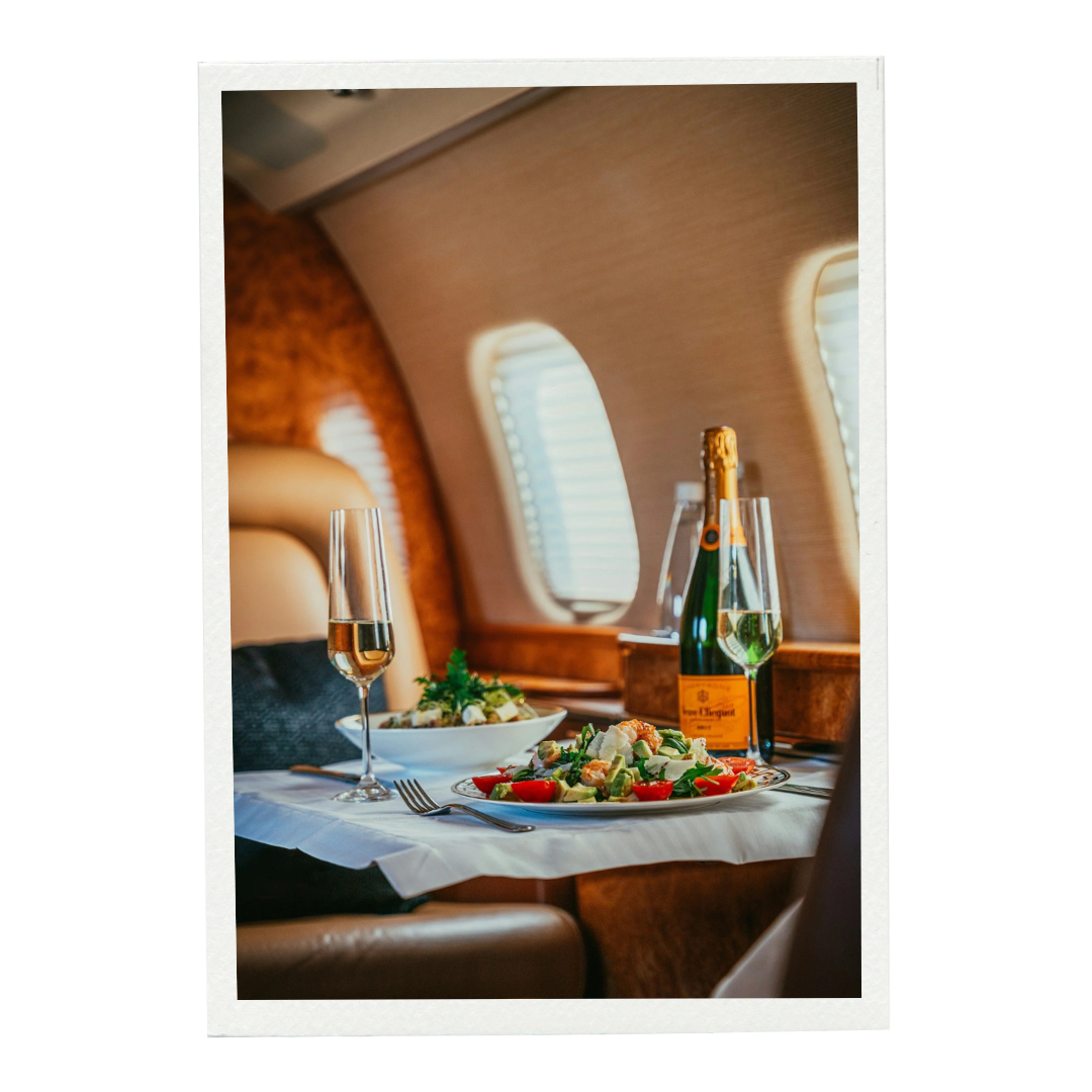 A luxurious airplane dinner setting with a glass of champagne, a bottle of champagne, and a salad with tomatoes and greens on a white tablecloth inside a plane with beige and wooden interior