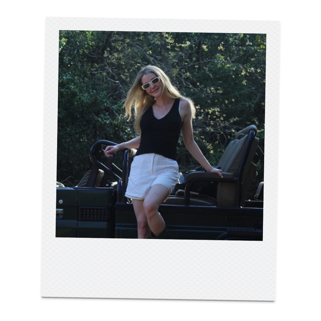 A woman with blonde hair wearing sunglasses, a black sleeveless top, and white shorts standing on a vehicle outdoors with trees in the background.