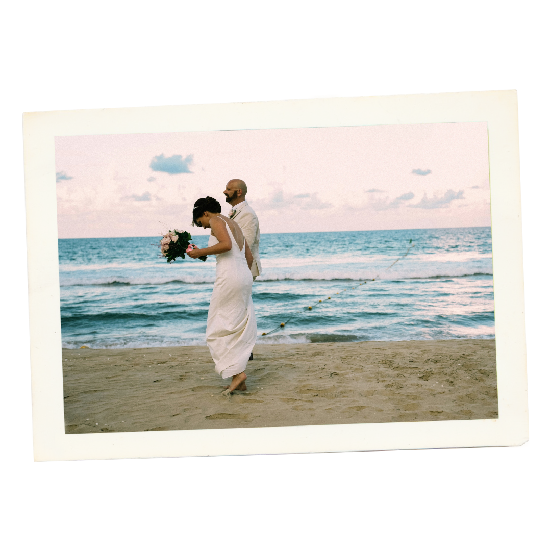 A couple walking on the beach during sunset, with the woman holding a bouquet of flowers, wearing a white wedding dress, and the man dressed in a white suit.