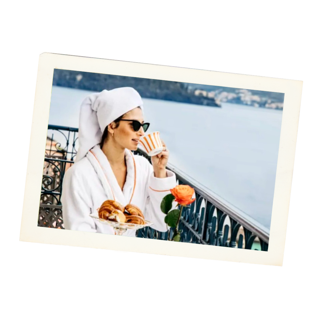 Woman in sunglasses and bathrobe with towel on head, drinking coffee on balcony with view of water, holding a plate of croissants and a rose.