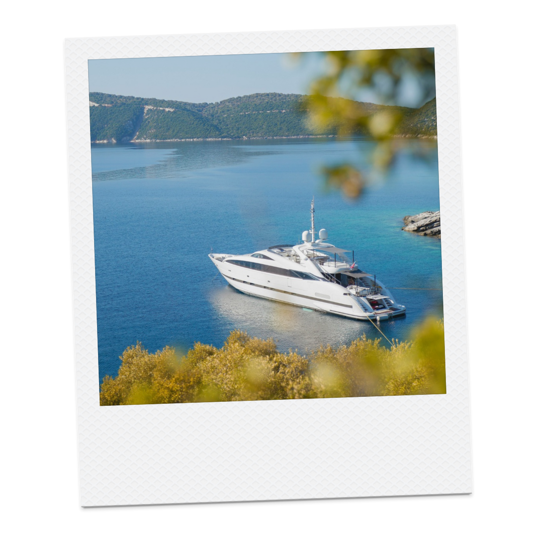 A white luxury yacht anchored near the shore in a calm, blue lake with forested hills in the background.