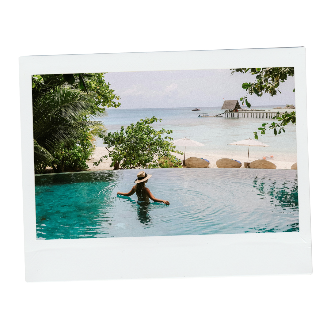 Woman in a hat relaxing in an infinity pool overlooking a beach with umbrellas, lounge chairs, and a pier extending into the ocean.