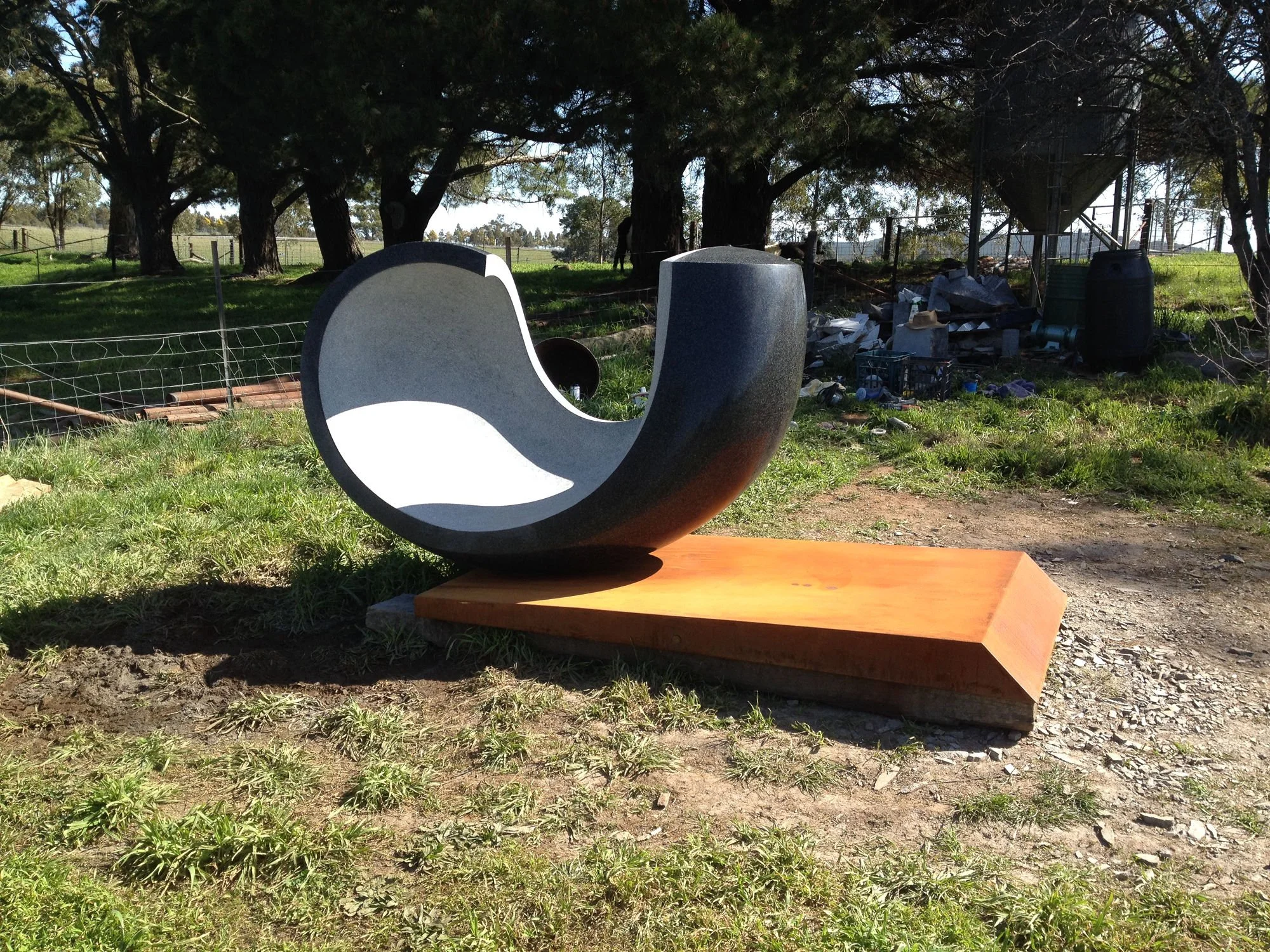 Outdoor scene with a modern sculpture in the foreground, trees providing shade, and a fence enclosing the area. There is a dirt ground and various objects scattered, including buckets and trash.