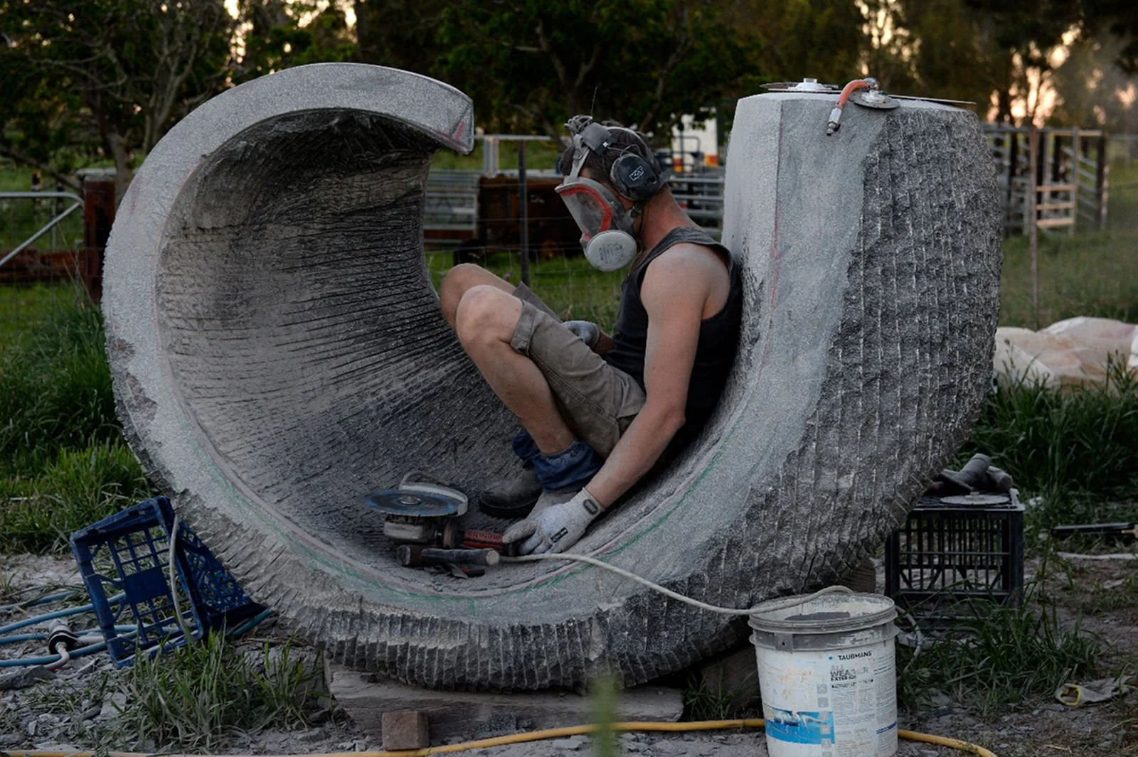 A person wearing protective gear, including a helmet and ear protection, is working on a large, hollow sculpture outdoors. The person is using a power tool, likely for grinding or cutting, with various tools and materials scattered around.