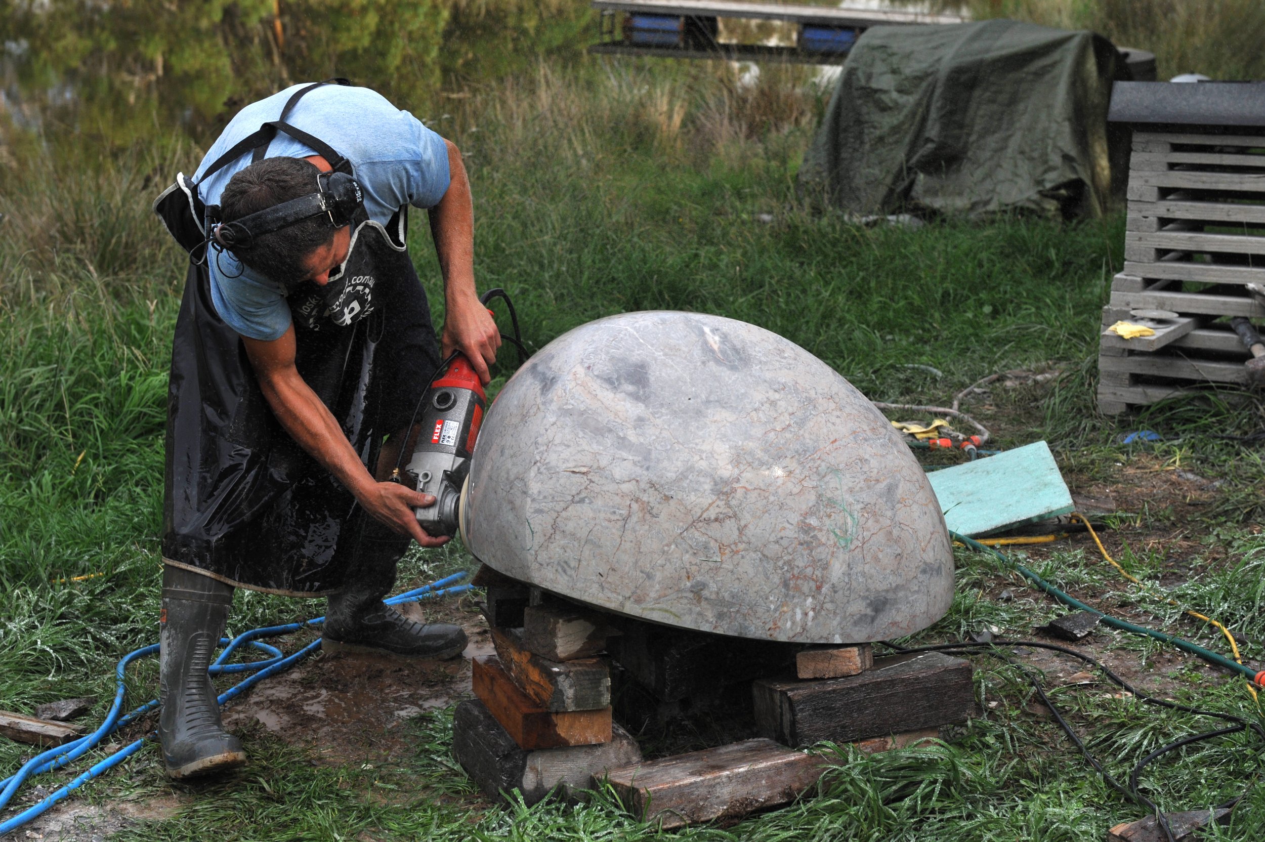 Person working outdoors with power tools, shaping a large stone or sculpture, nearby tools and materials on grass and wooden supports.