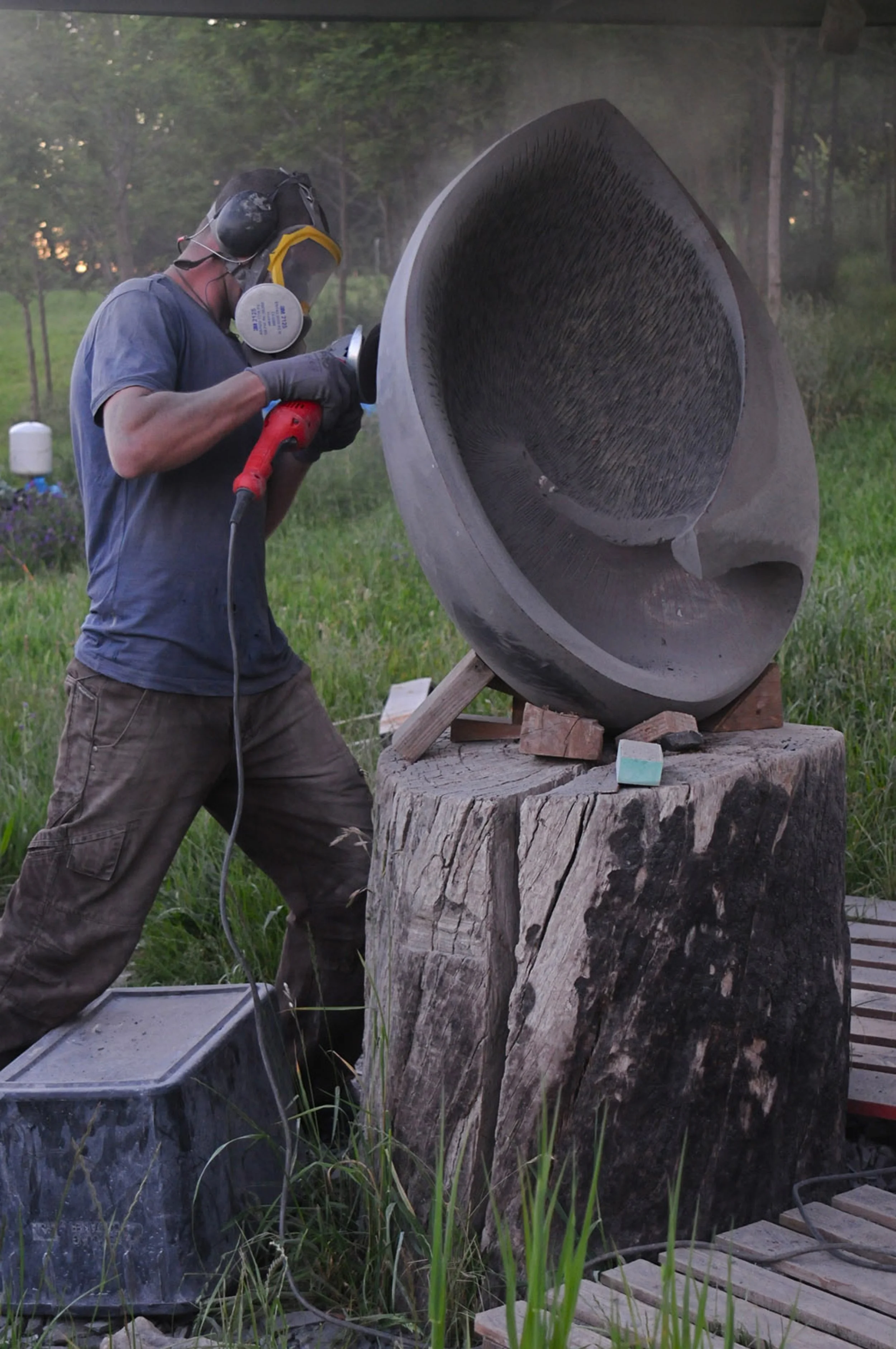 A man wearing safety gear, including a helmet, face mask, and gloves, is using an angle grinder on a large hollowed sculpture
 outdoors. The sculpture is placed on a sturdy wooden stand, and there are trees and greenery in the background.
