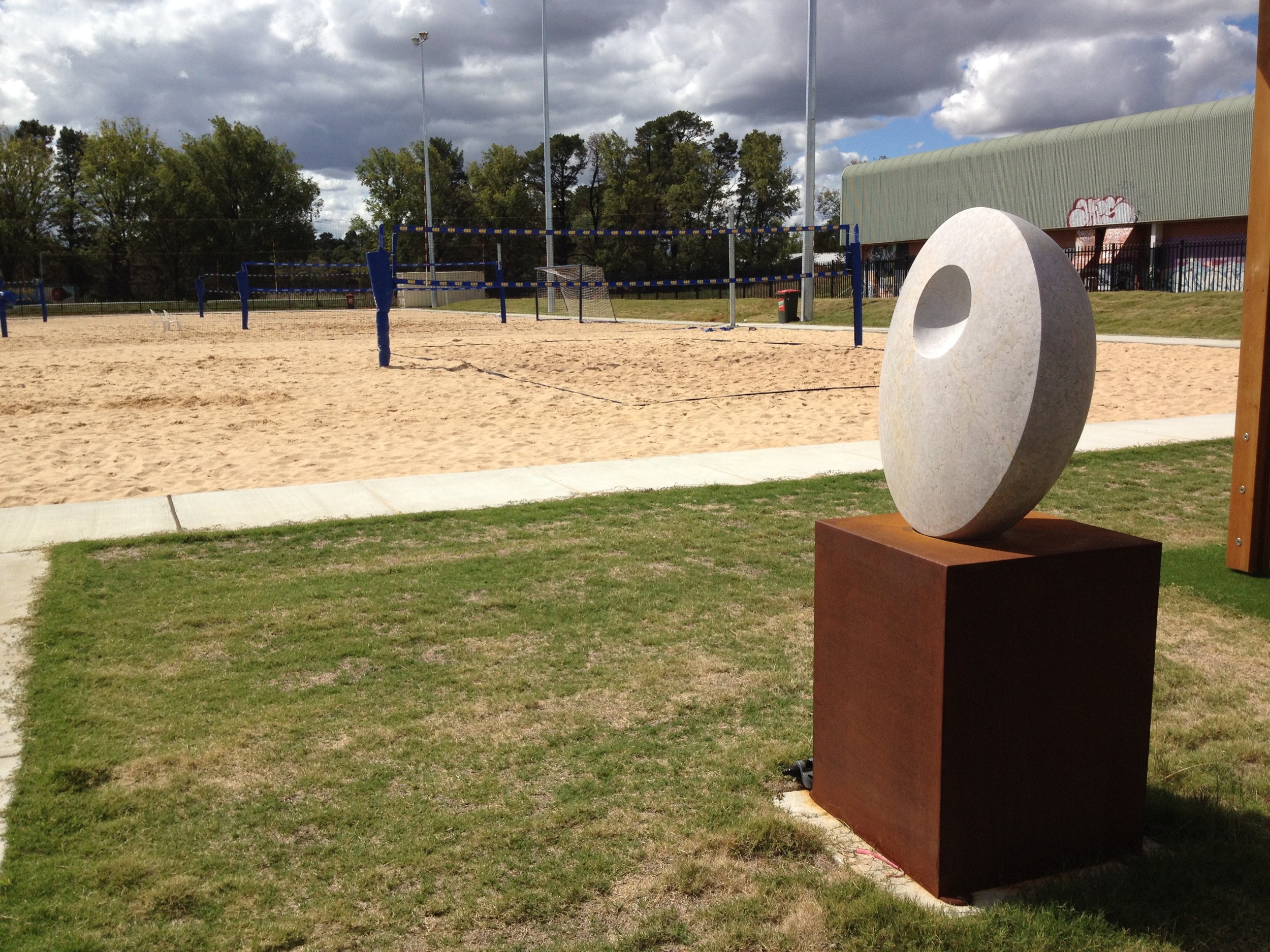 An outdoor sand volleyball court with blue net, surrounded by trees and a fence, under a partly cloudy sky, with a large abstract sculpture in the foreground.