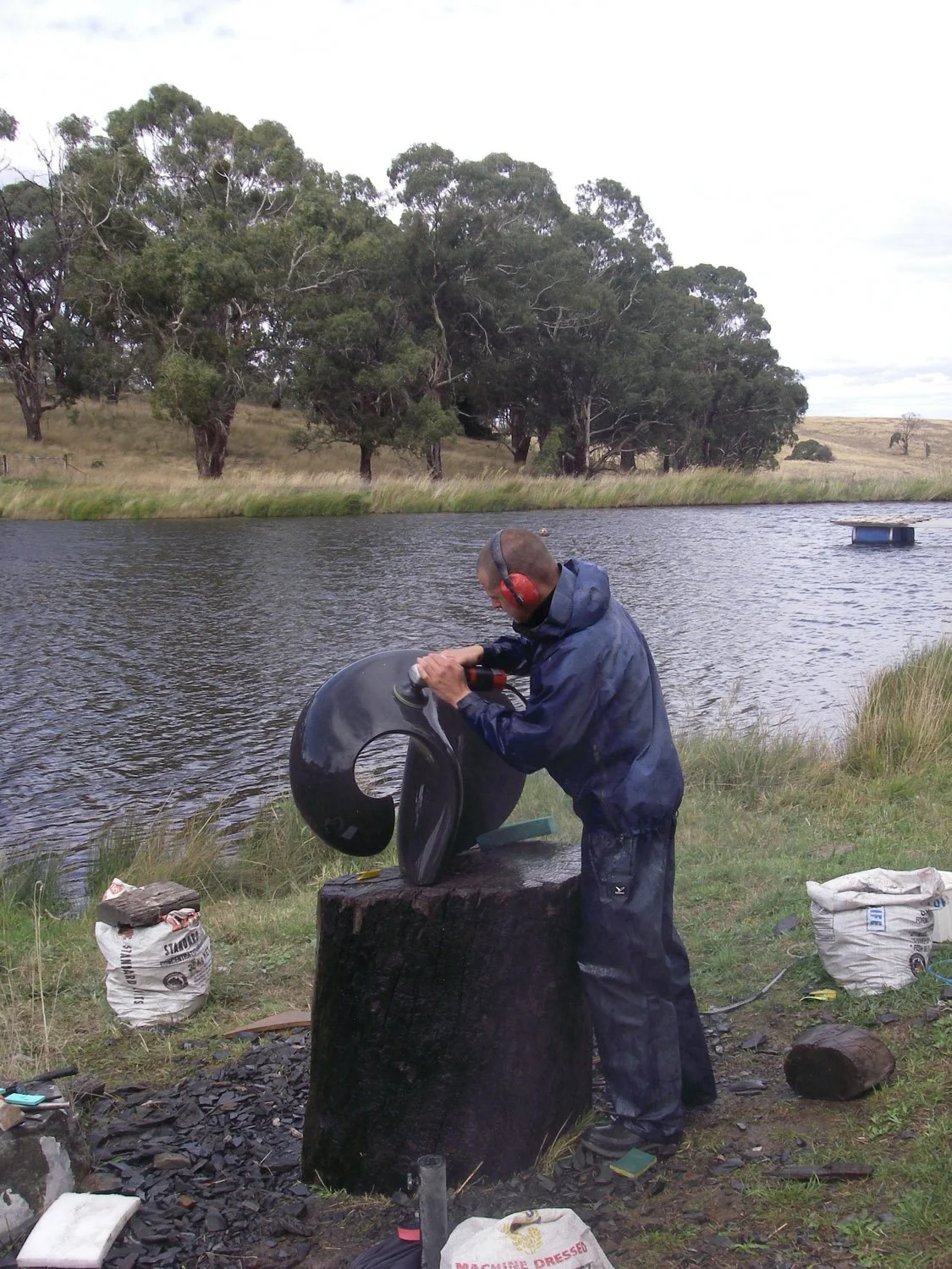 A man wearing headphones and waterproof clothing is carving a black sculpture with a power tool by a lake, surrounded by grass and trees.