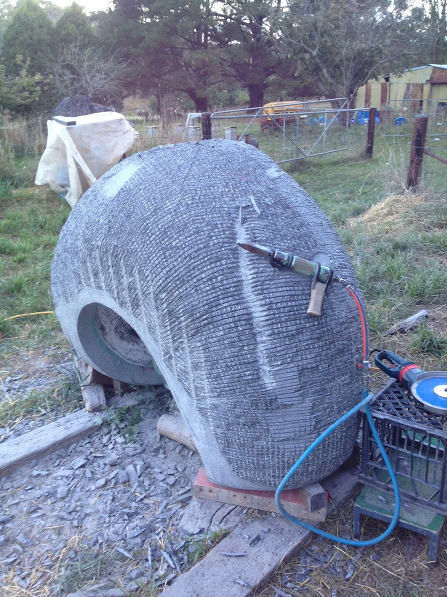 A large, cylindrical sculpture being cut or planed with a power tool in an outdoor setting, with tools, debris, and a fence in the background.