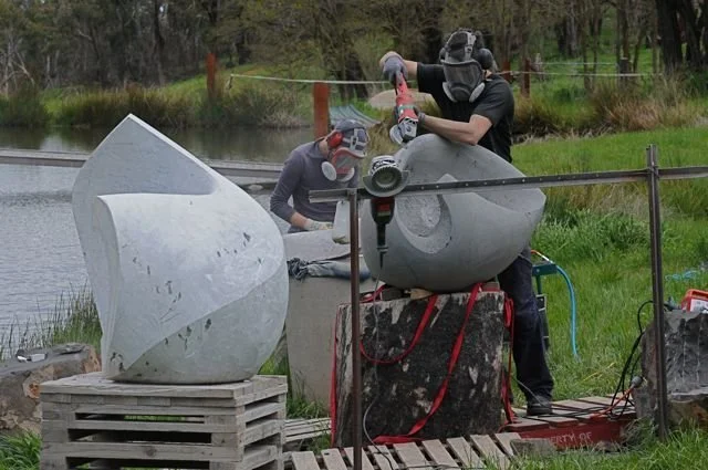 Two artists working on sculptures made of grey stone outdoors near a pond, with trees and grass in the background.