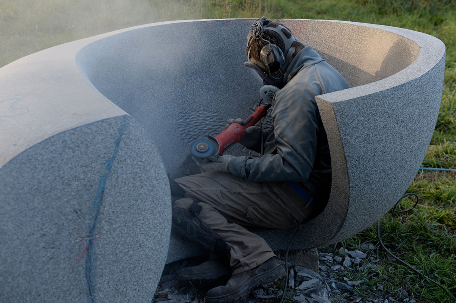 A worker in safety gear using a power tool inside a large stone sculpture outdoors.