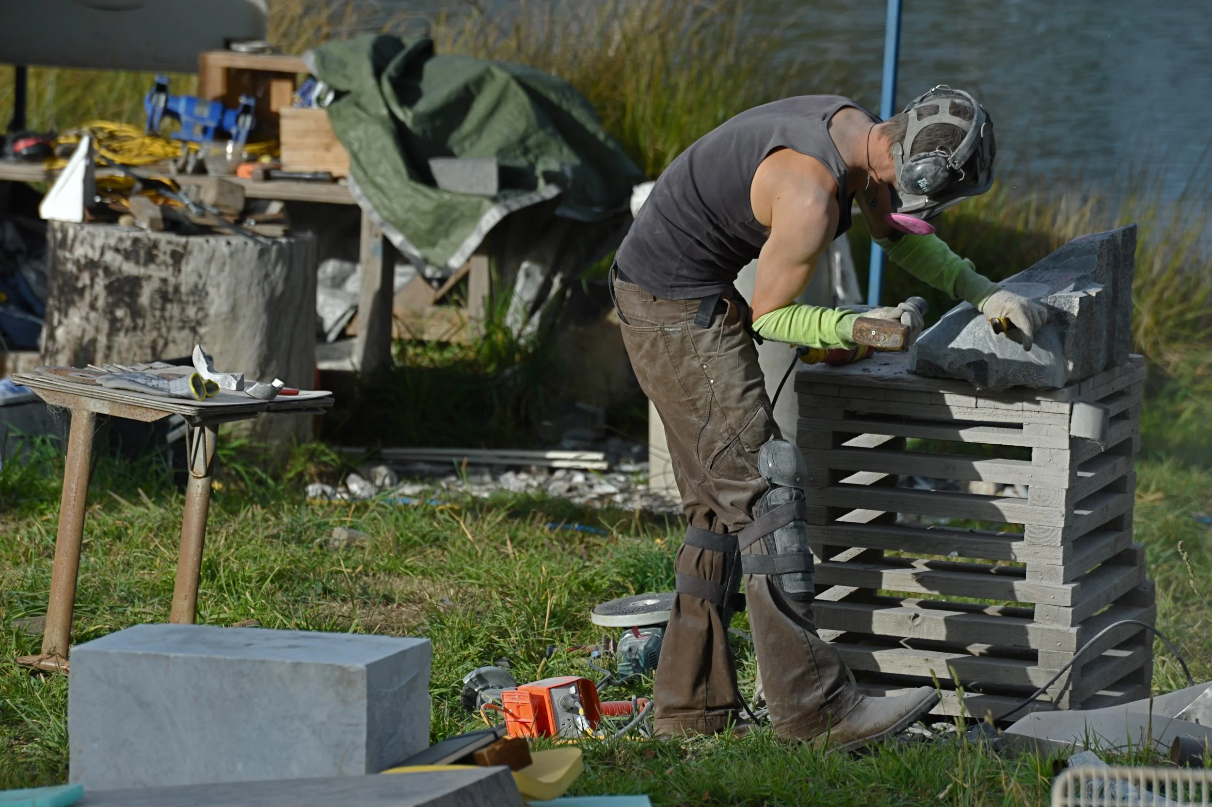 A person working outdoors near a body of water, wearing safety gear and working on a stone sculpture, with tools and supplies scattered around.