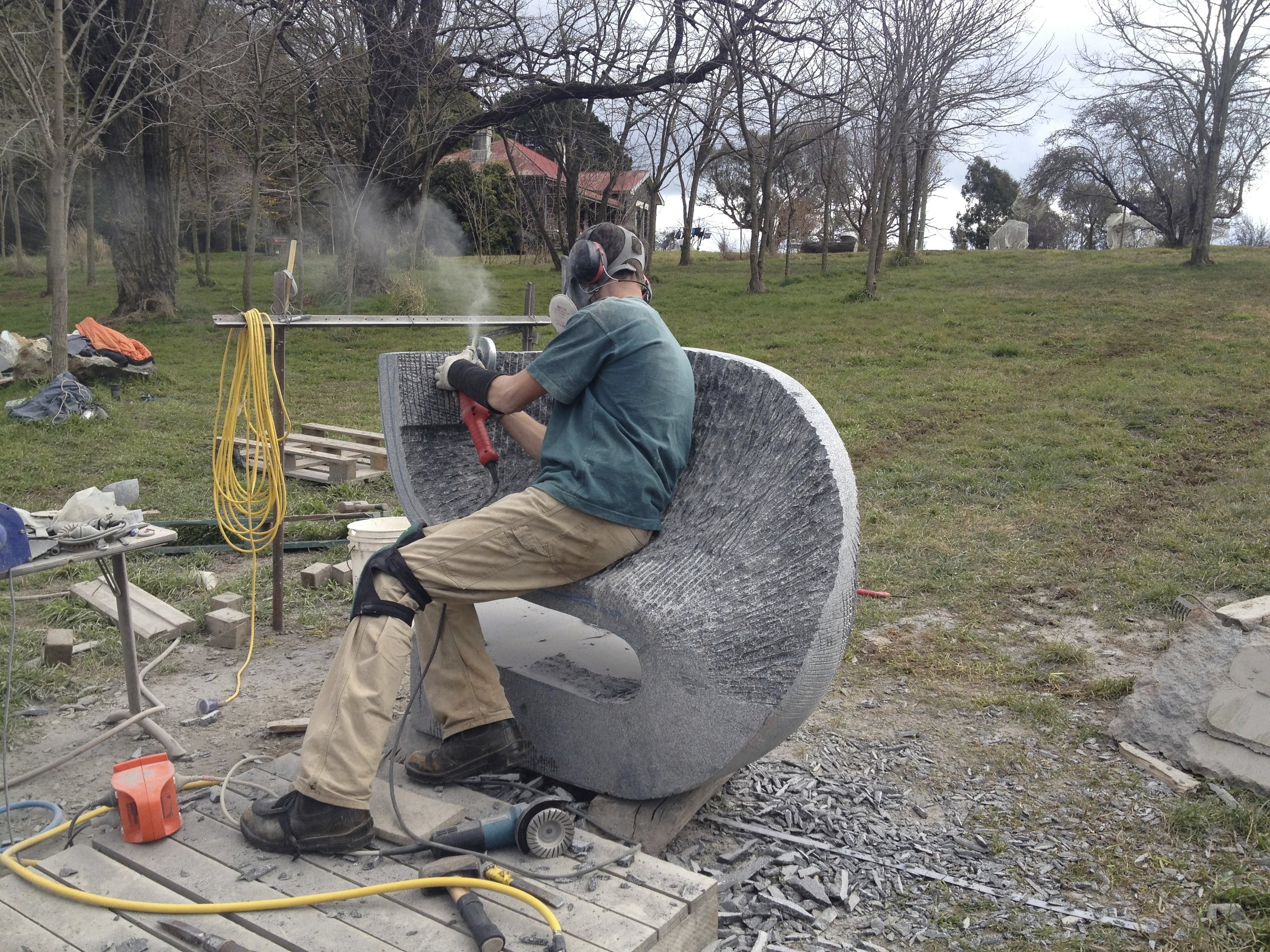 A person wearing safety gear working on what appears to be a large stone sculpture outdoors. The person is using power tools and sitting on a curved stone piece surrounded by various tools and equipment. The scene takes place in an open grassy area w