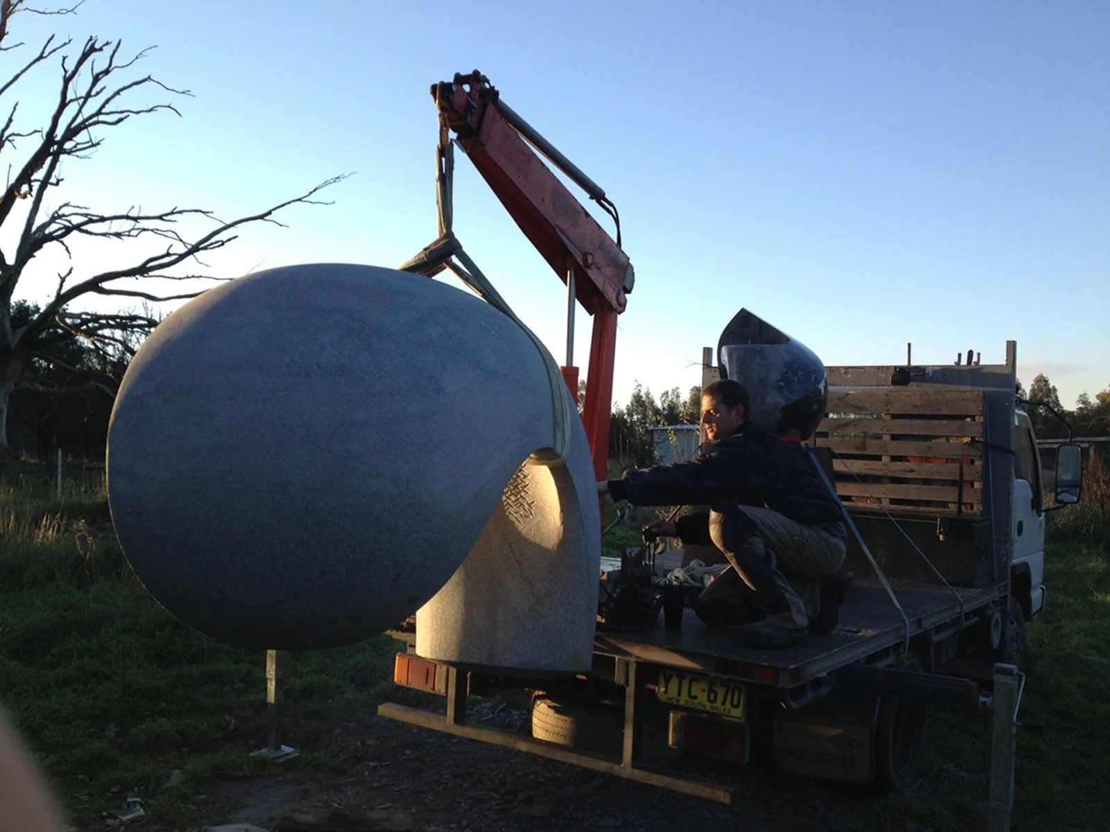 Man crouching on the back of a pickup truck, working on a large outdoor sculpture resembling a sphere or planet with a textured surface, in a rural setting during dusk.