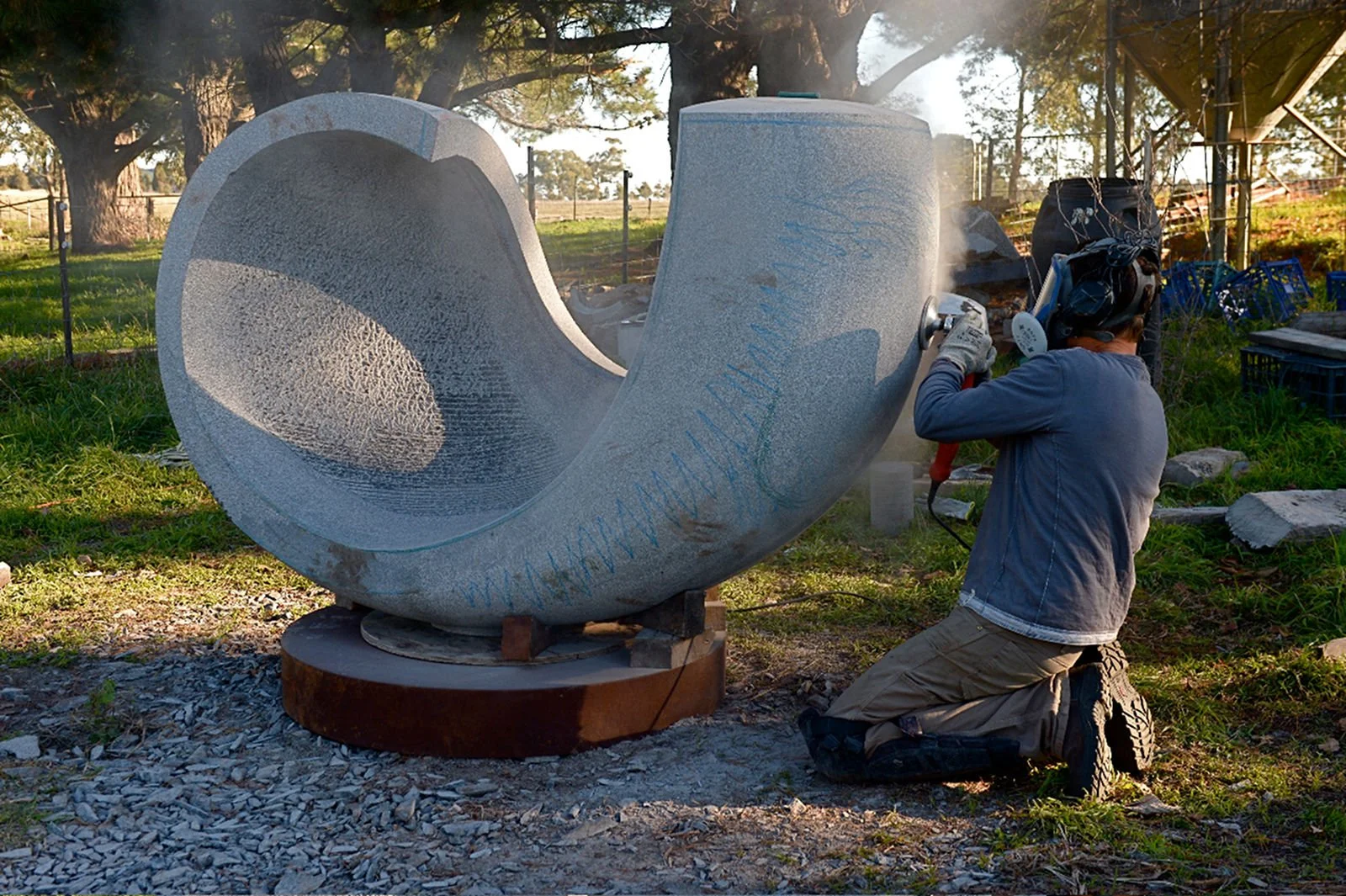 A person wearing a helmet and gloves is using a power tool to carve or drill into a large, hollow, gray stone sculpture in a grassy outdoor area with trees in the background.