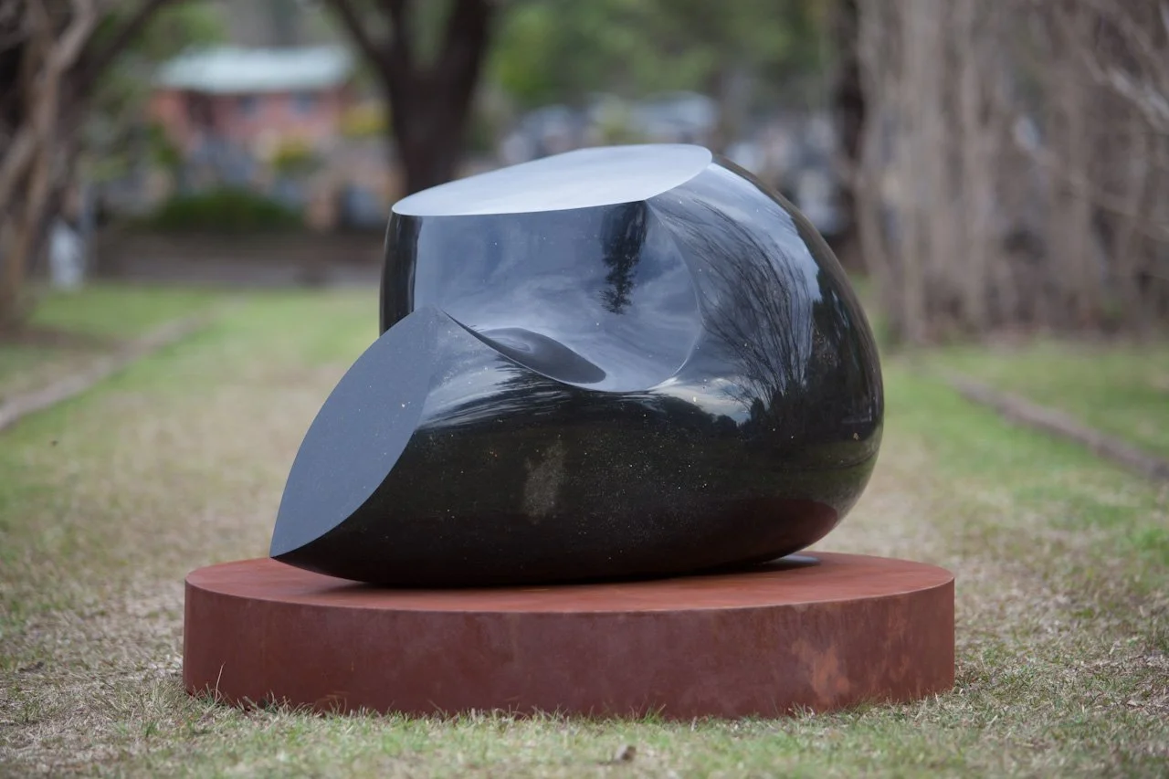 A polished black sculpture on a round, rust-colored pedestal outdoors with trees and buildings in the background.
