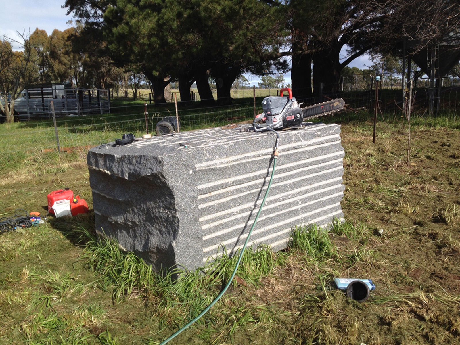 A large rectangular granite stone with horizontal grooves on its surface, placed outdoors on grassy ground. Construction tools and equipment are positioned on and around the stone, including a chainsaw on top, a red fuel container, and other small it