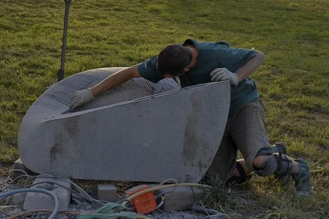 Person working on large sculpture outdoors, kneeling and examining it closely.