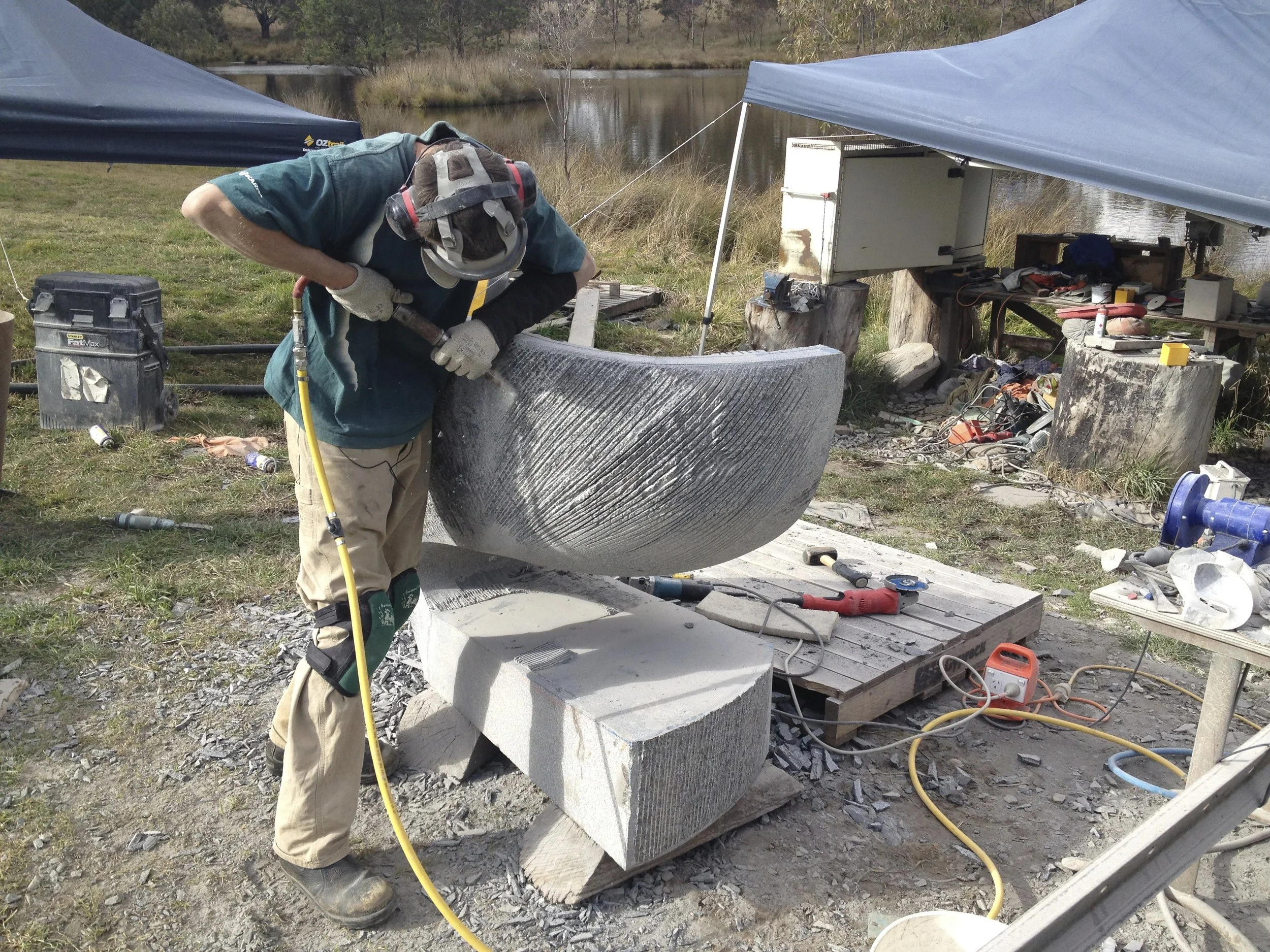 A person wearing safety goggles and gloves is carving or polishing a large, rounded stone or marble block outdoors near a body of water, with tents and tools scattered around.