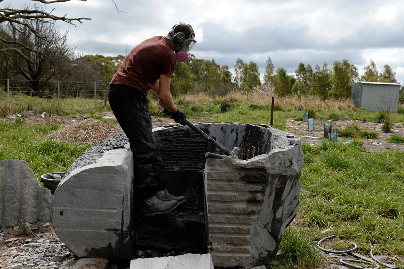 A man wearing a mask, gloves, and protective glasses is working on a large stone sculpture outdoors, using a hammer and chisel to carve the stone. The setting is a grassy area with trees and a metal shed in the background.