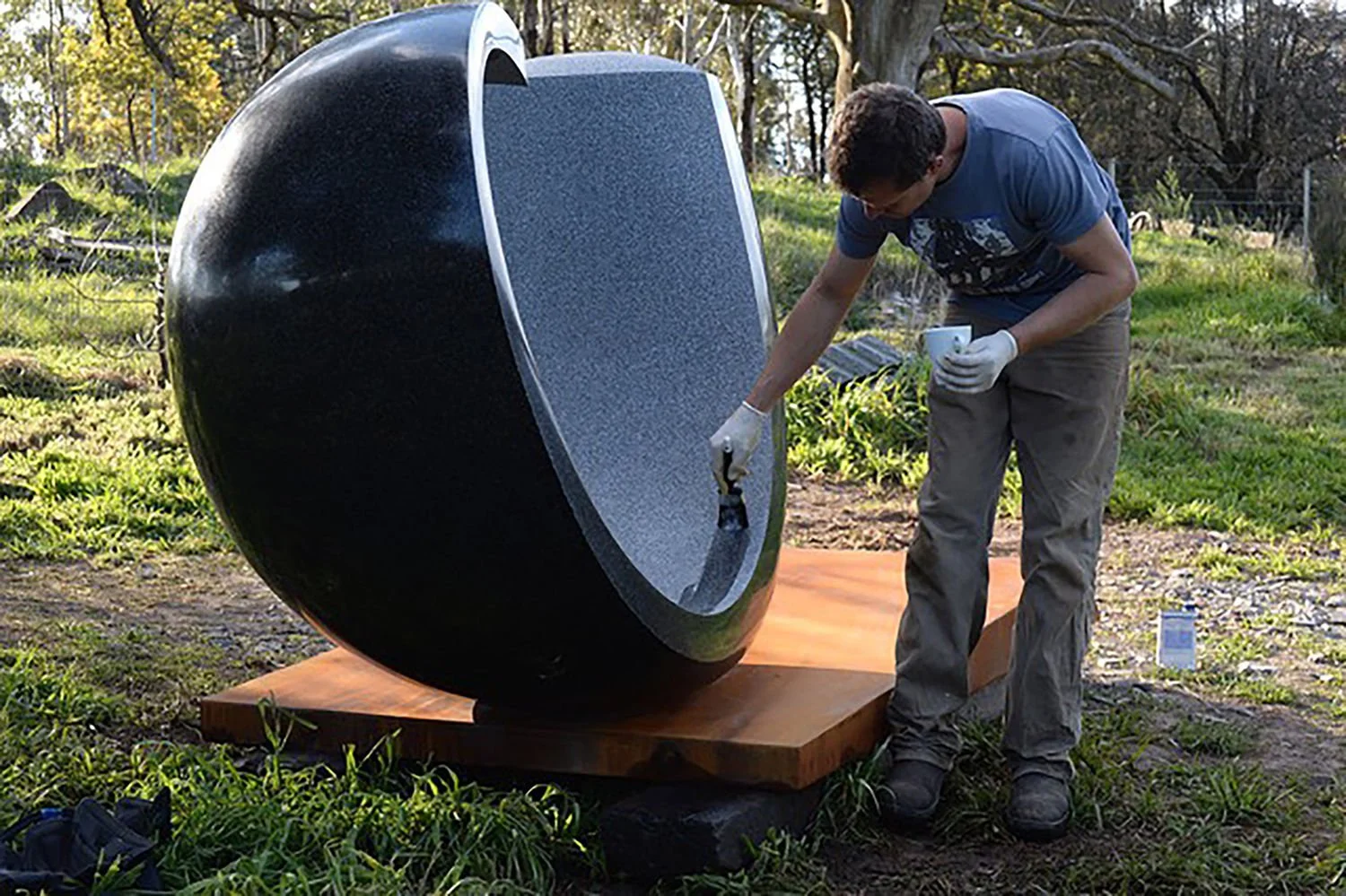A person working on a large, oval-shaped, black and gray object outdoors, wearing gloves and holding tools, with trees and grass in the background.