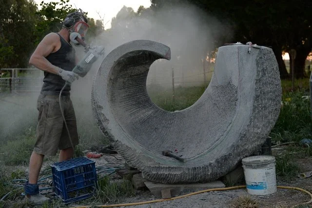 A person wearing a mask and gloves is working on a large granite sculpture shaped like a crescent moon, outdoors with trees and a fence in the background.