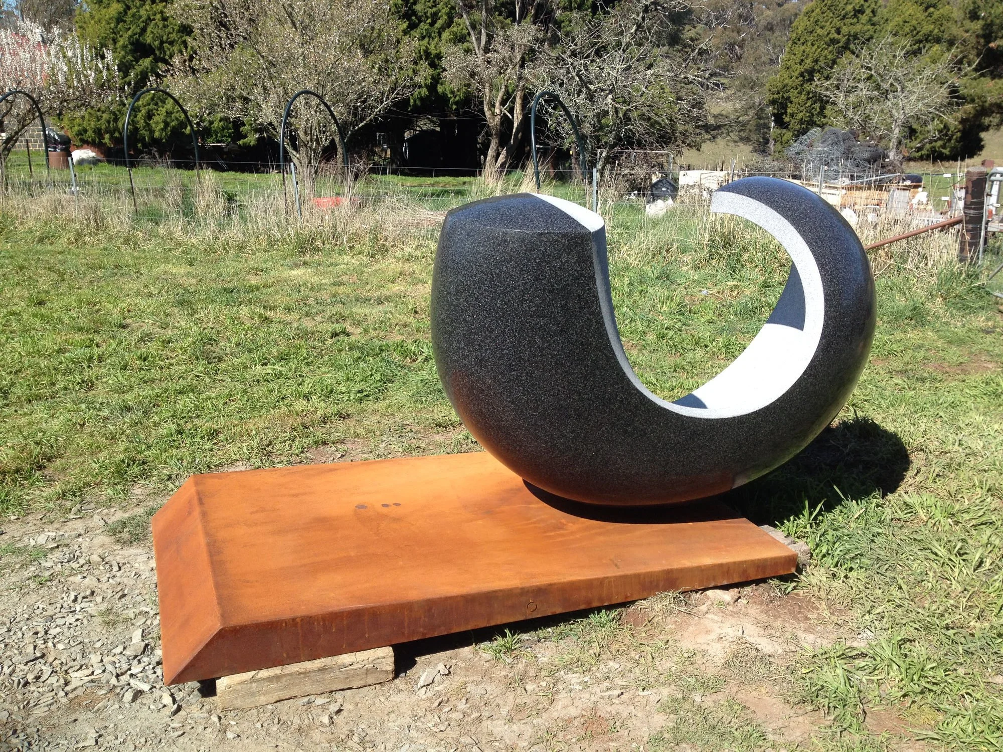 A modern black and white sculpture with a circular hollow center, placed on a metal platform outdoors with grass, trees, and fencing in the background.