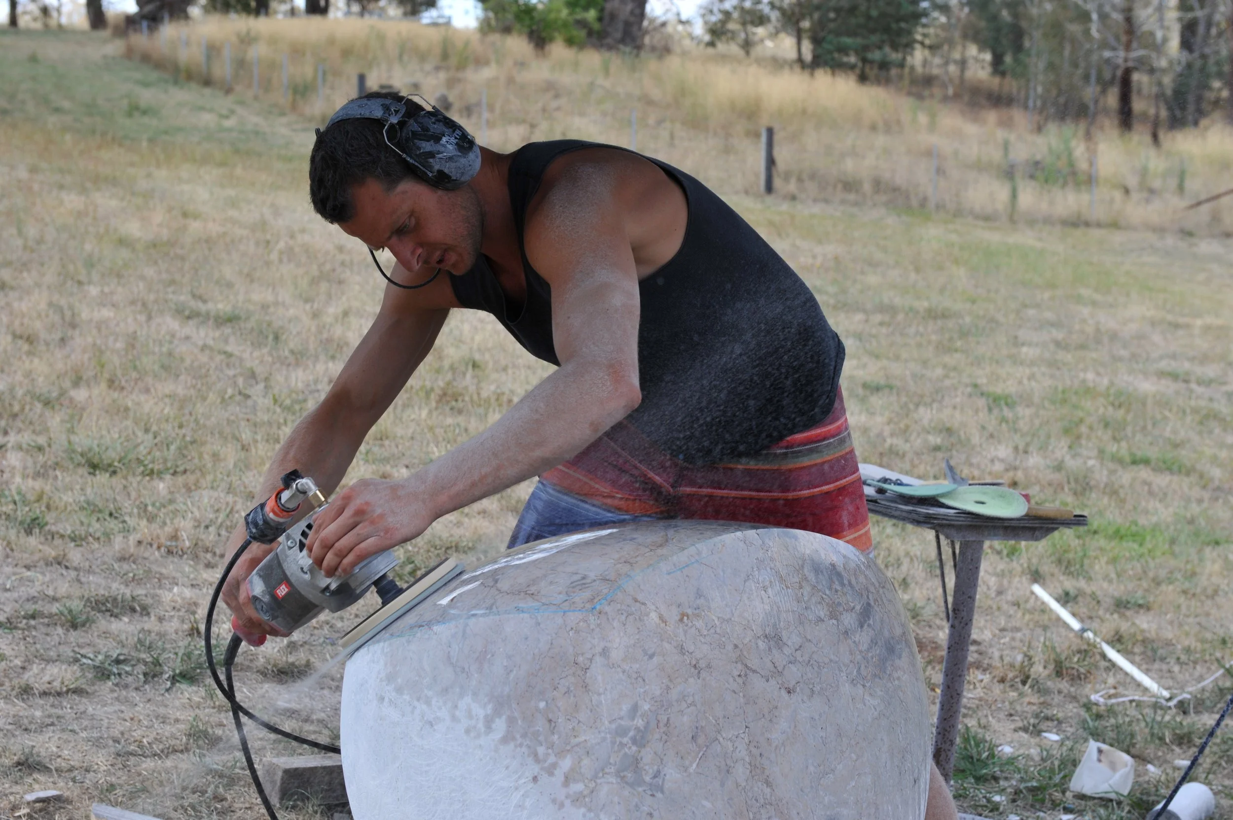 A man working with power tools on a large spherical object outdoors in a grassy field.